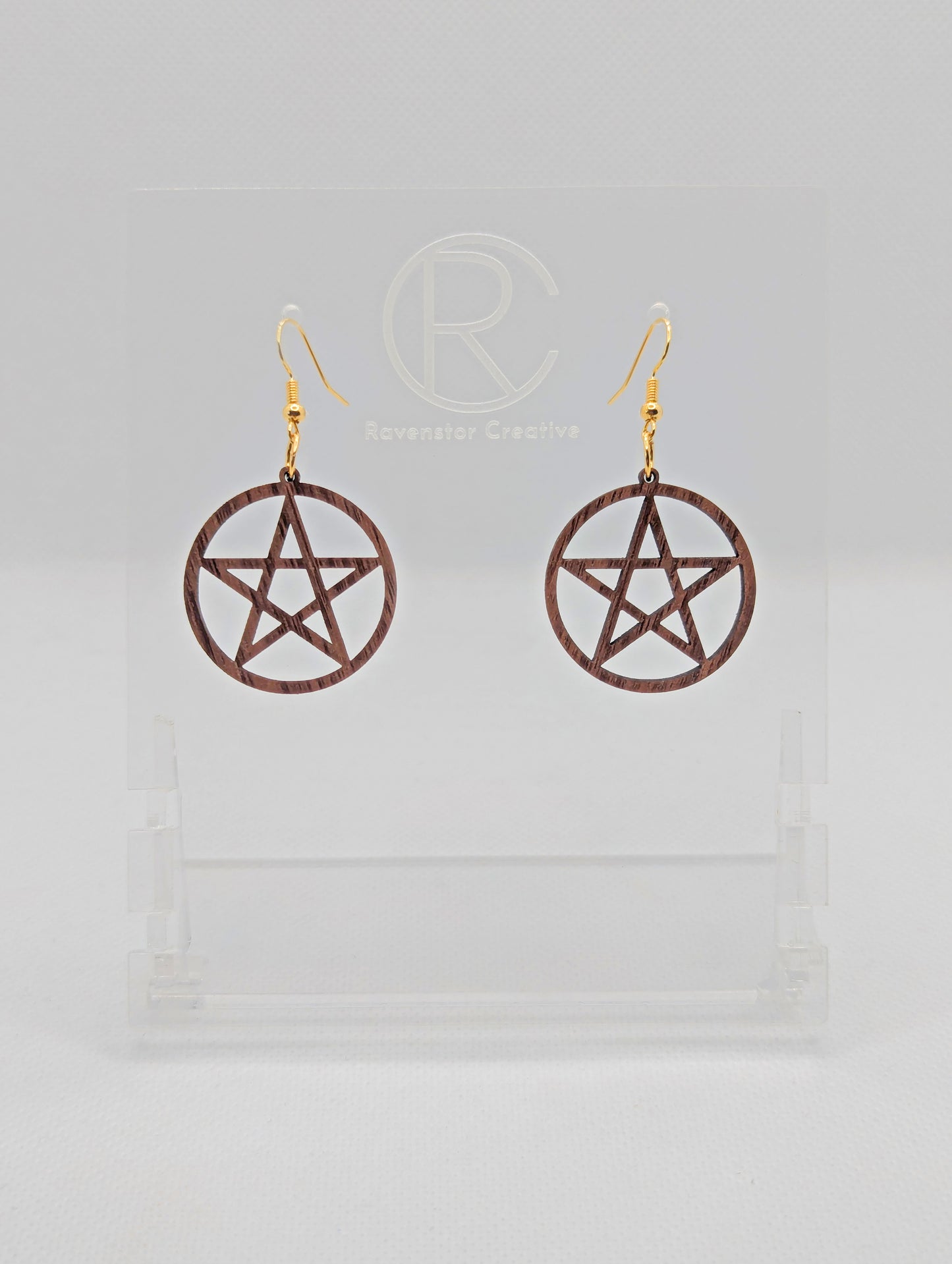 Shows a pair of earrings with gold ear wire hook fixings. They are Pentacle / Pentagram Symbols. The colour of them are Walnut Wood acrylic. They on a see through stand that has the Ravenstor Creative Logo and its against a white background.