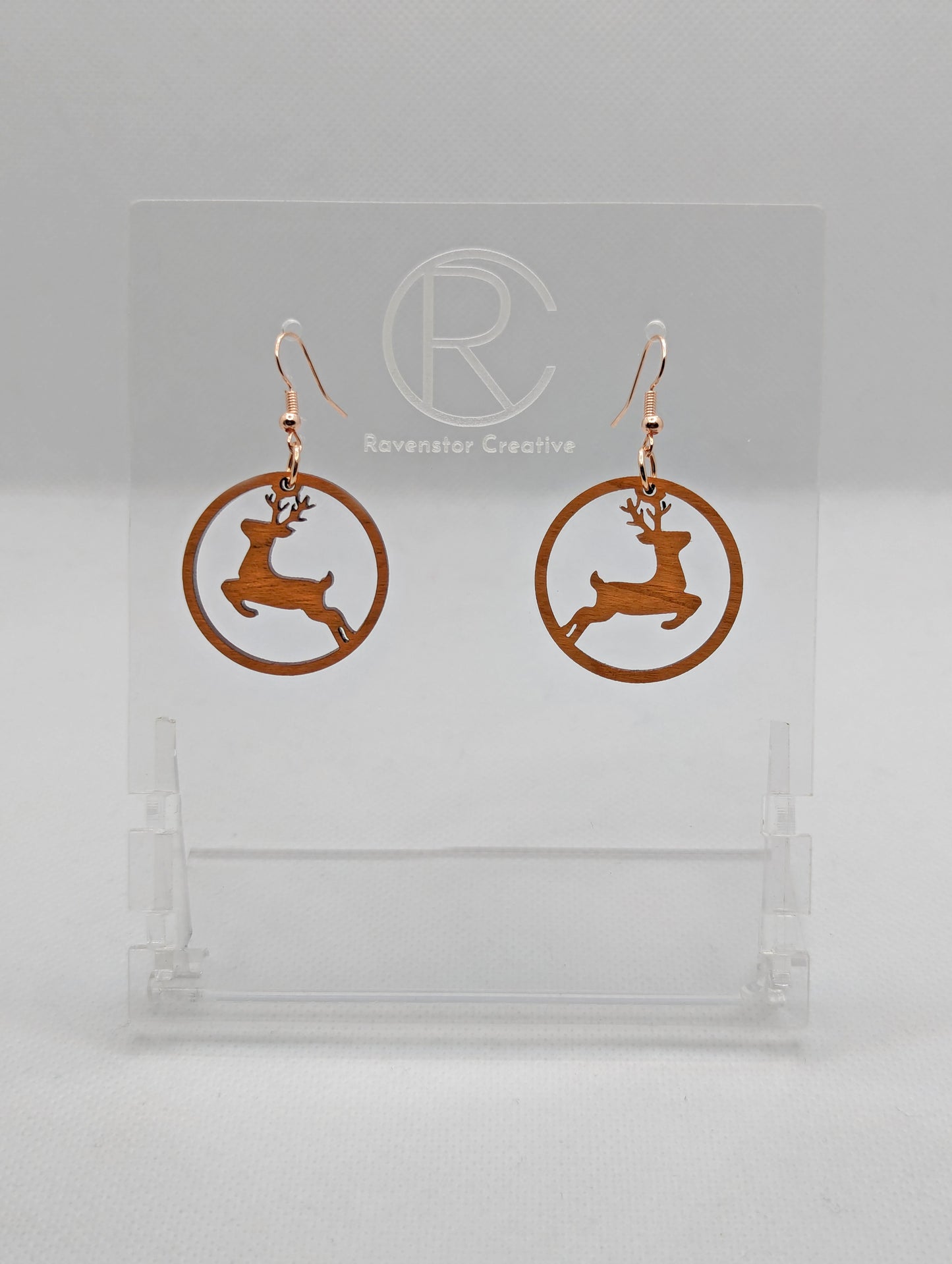 Shows a pair of earrings that have rose gold ear wire hook fixings. They are circles with prancing reindeer/deer cut out. They are in Cherry Wood and are against a white background on see through stand with the Ravenstor Creative logo on it.