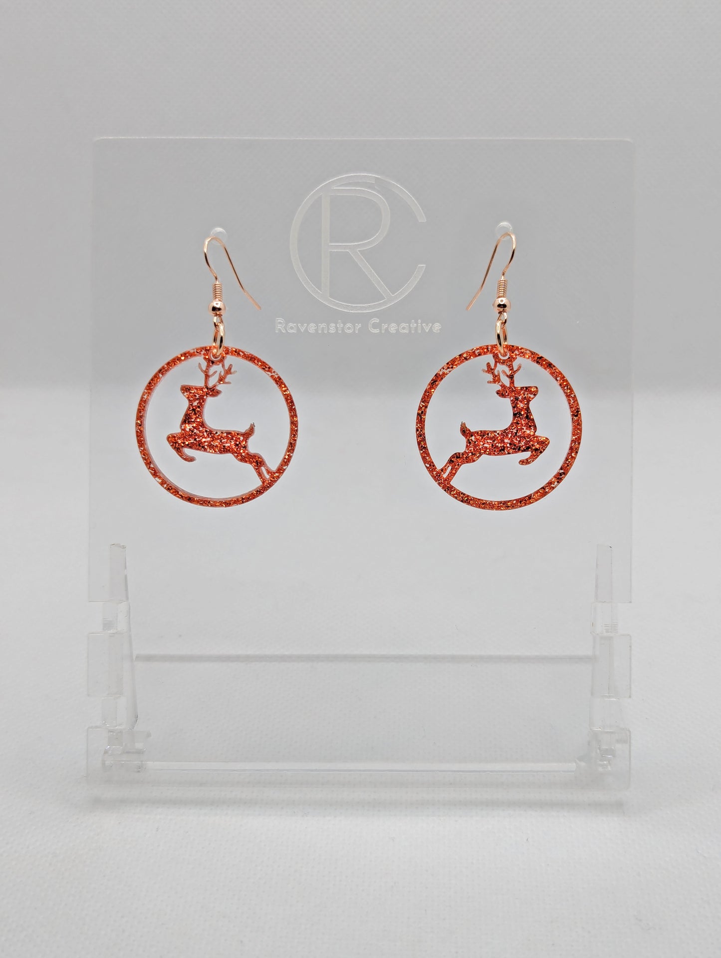 Shows a pair of earrings that have rose gold ear wire hook fixings. They are circles with prancing reindeer/deer cut out. They are in Copper Glitter and are against a white background on see through stand with the Ravenstor Creative logo on it.