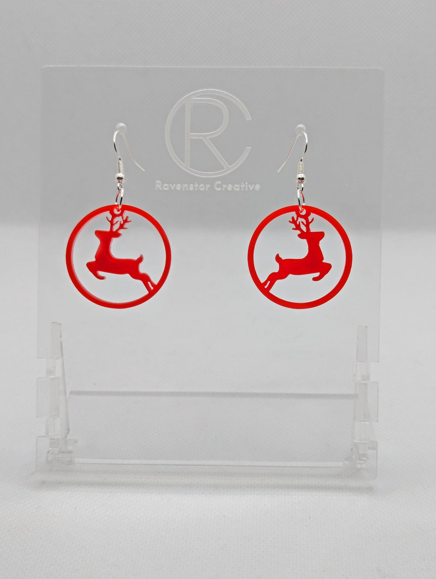 Shows a pair of earrings that have silver ear wire hook fixings. They are circles with prancing reindeer/deer cut out. They are in Red Acrylic and are against a white background on see through stand with the Ravenstor Creative logo on it.