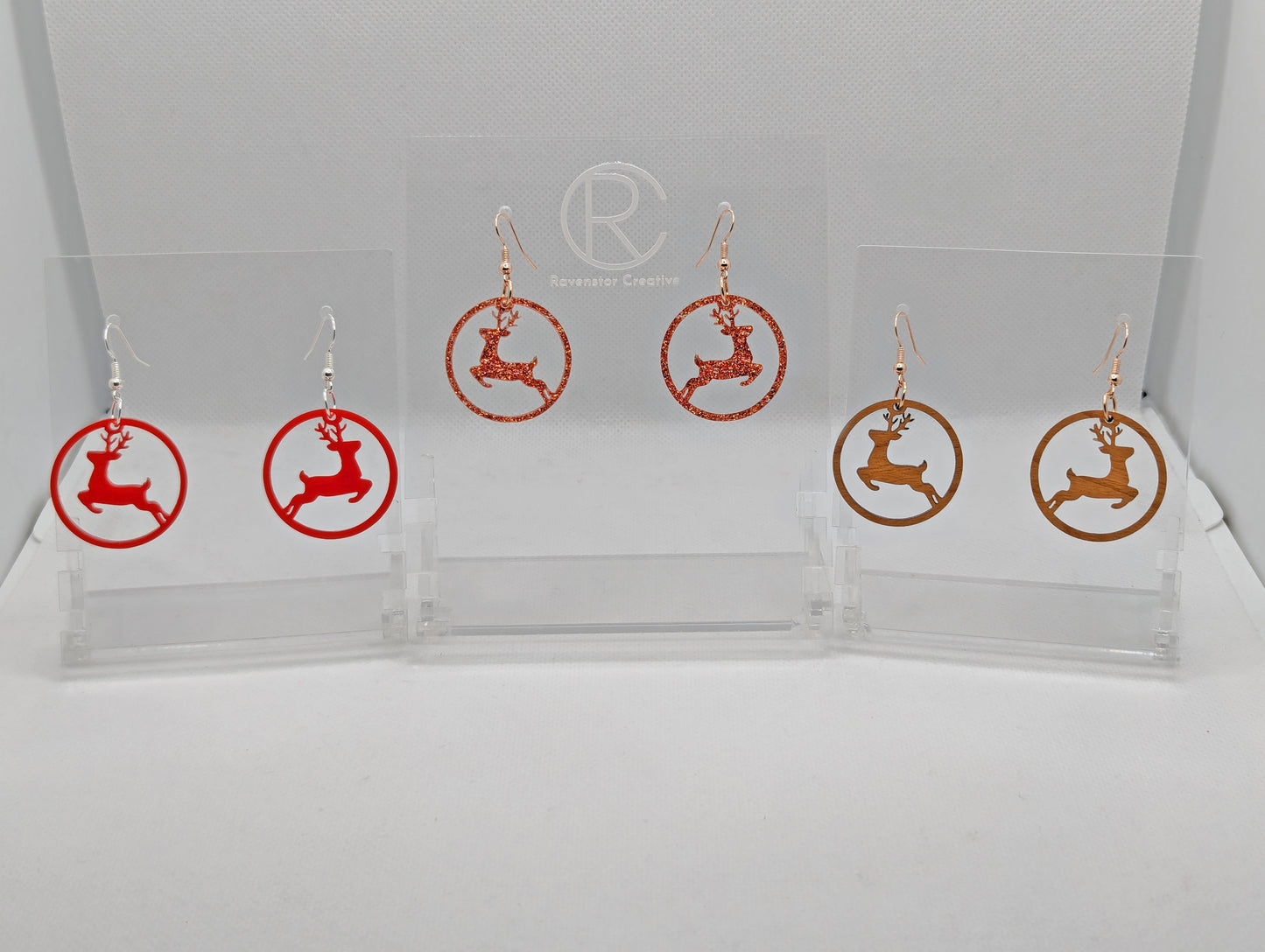 Shows three pairs of earrings that have ear wire hook fixings. They are a circle with prancing reindeer/deer cut out. The colours are cherry wood, Red and Copper Glitter. They are against a white background on see through stands.