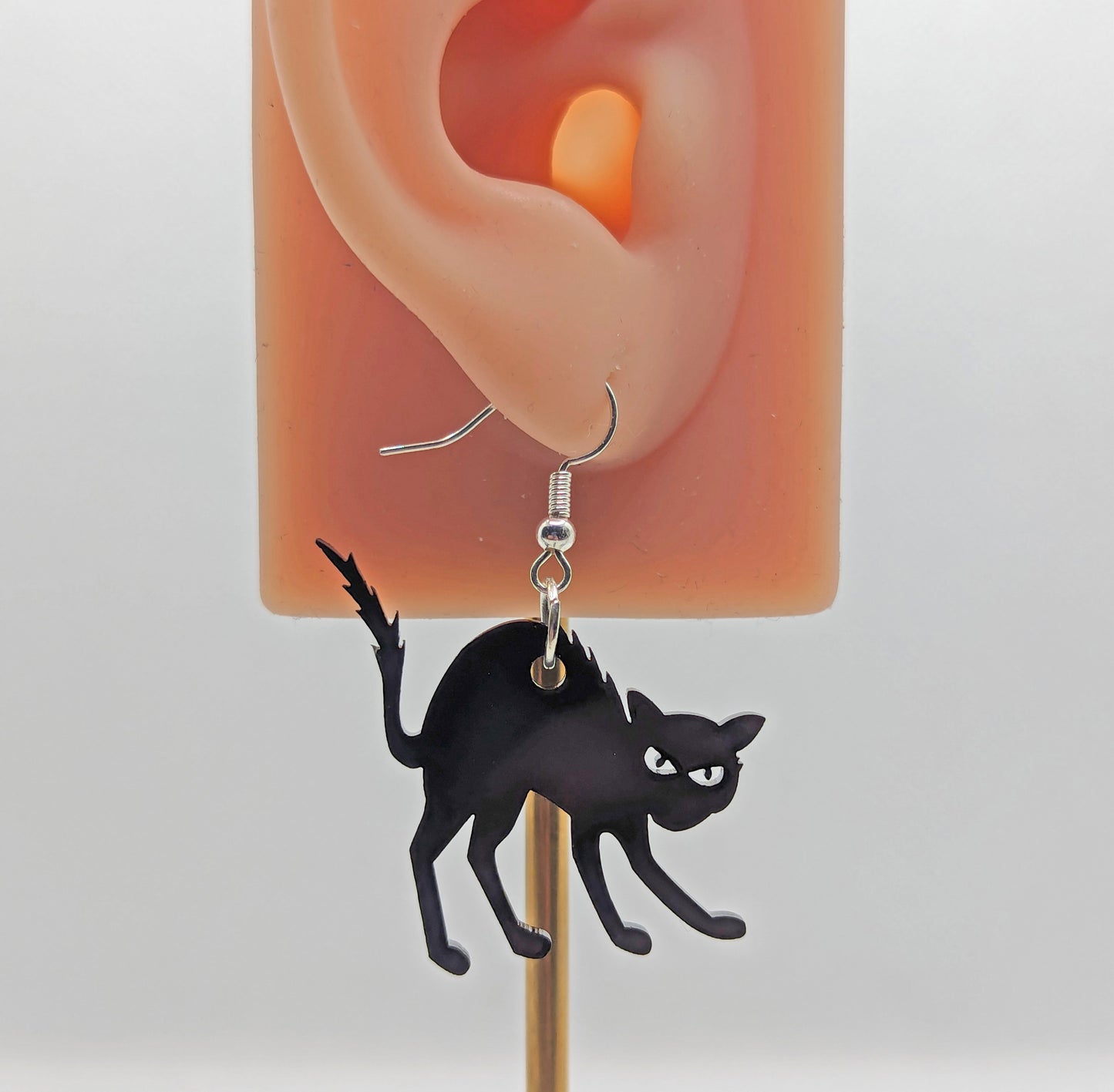 An earring on an ear to show the size with silver ear wire hook fixings. It is an angry or scared looking cat in black acrylic with thier heckles and tails up and fir on end against a white background.