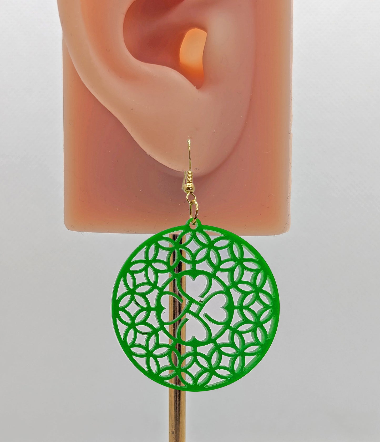 Shows an acrylic earring that has silver ear wire hook fixings. It is a circle with a shamrock four leaf clover design. Is it on an ear showing the size against a white background.
