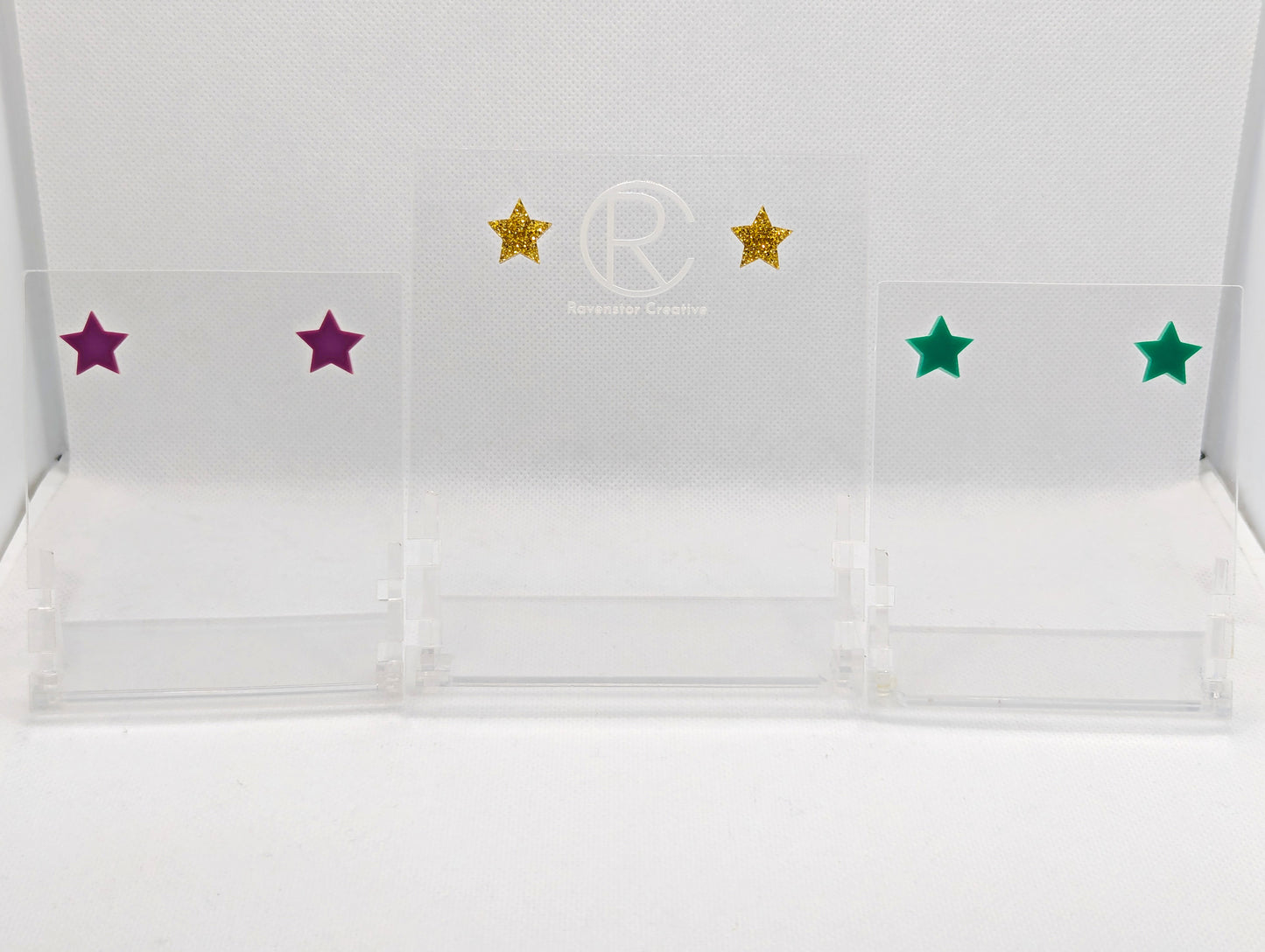 Shows three pairs of small acrylic studded earrings that are Stars. The colours are Grape, Gold Glitter and Grape. They are against a white background on a clear stand with the Ravenstor Creative logo on it.