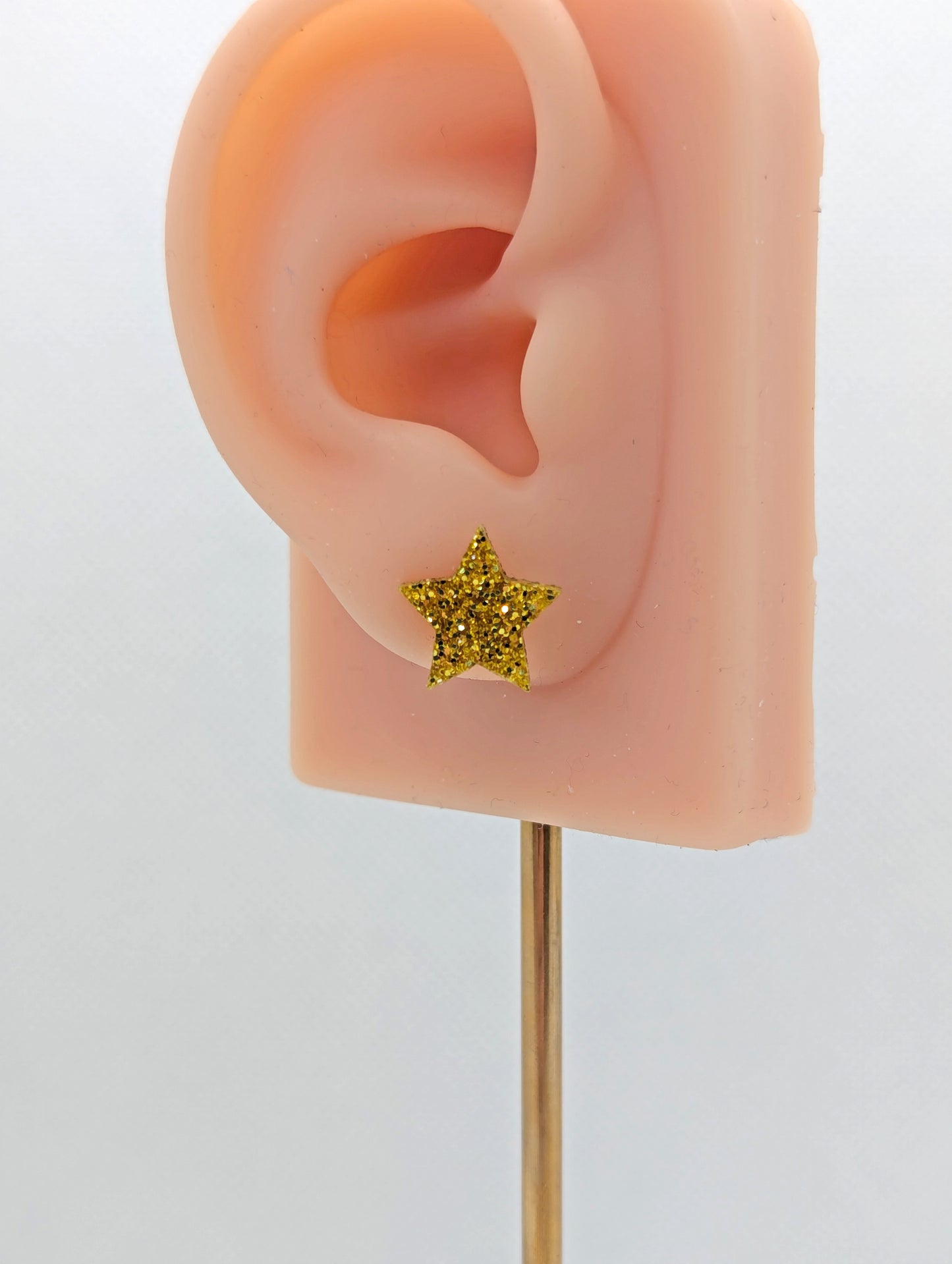 Shows a pair of small studded acrylic earrings that are Stars. They are Gold Glitter in colour. They are on an ear to show the size.