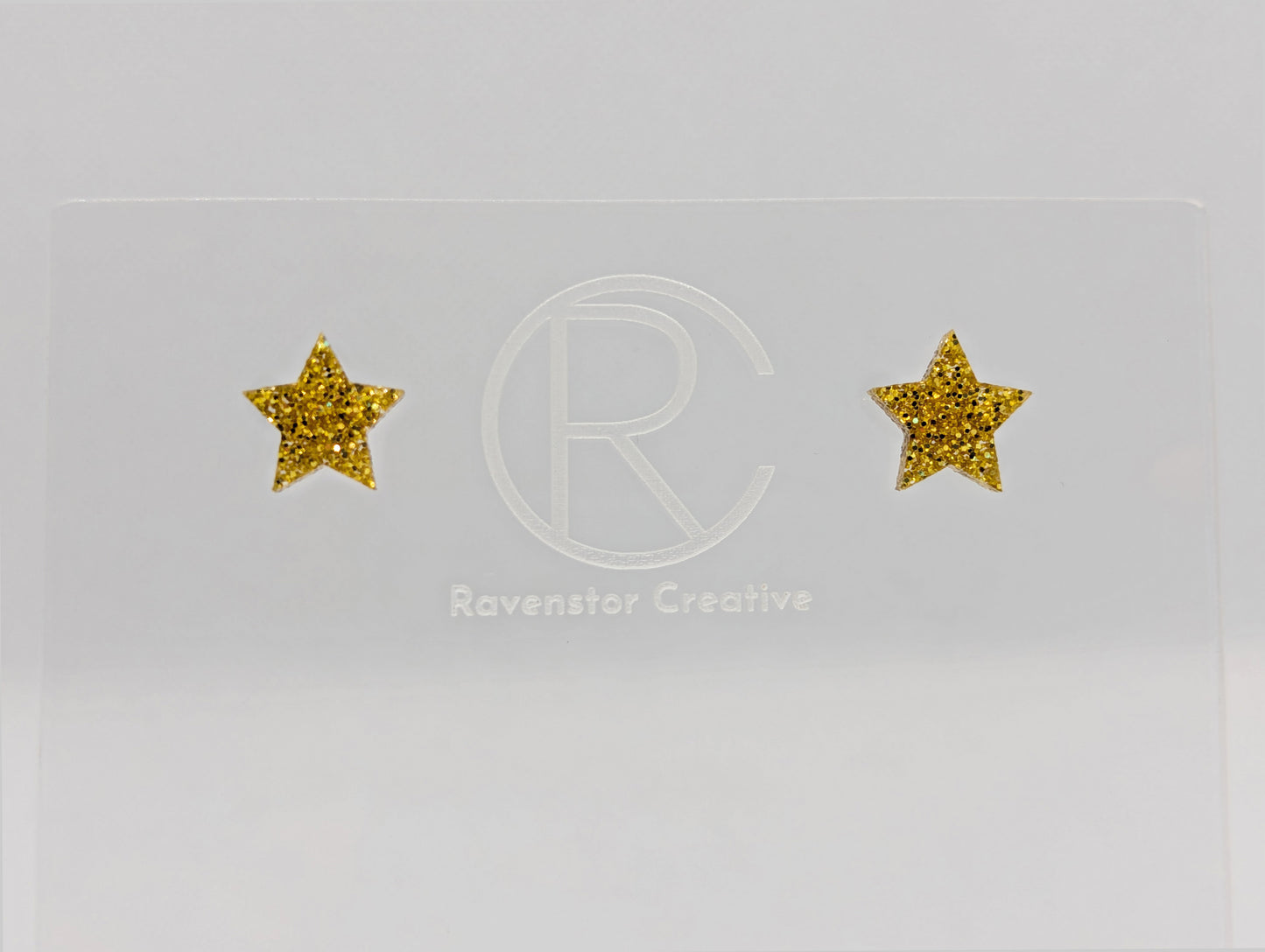 Shows a pair of small studded acrylic earrings that are Stars. They are Gold Glitter in colour. They are against a white background on see through stand with the ravenstor creative logo on.