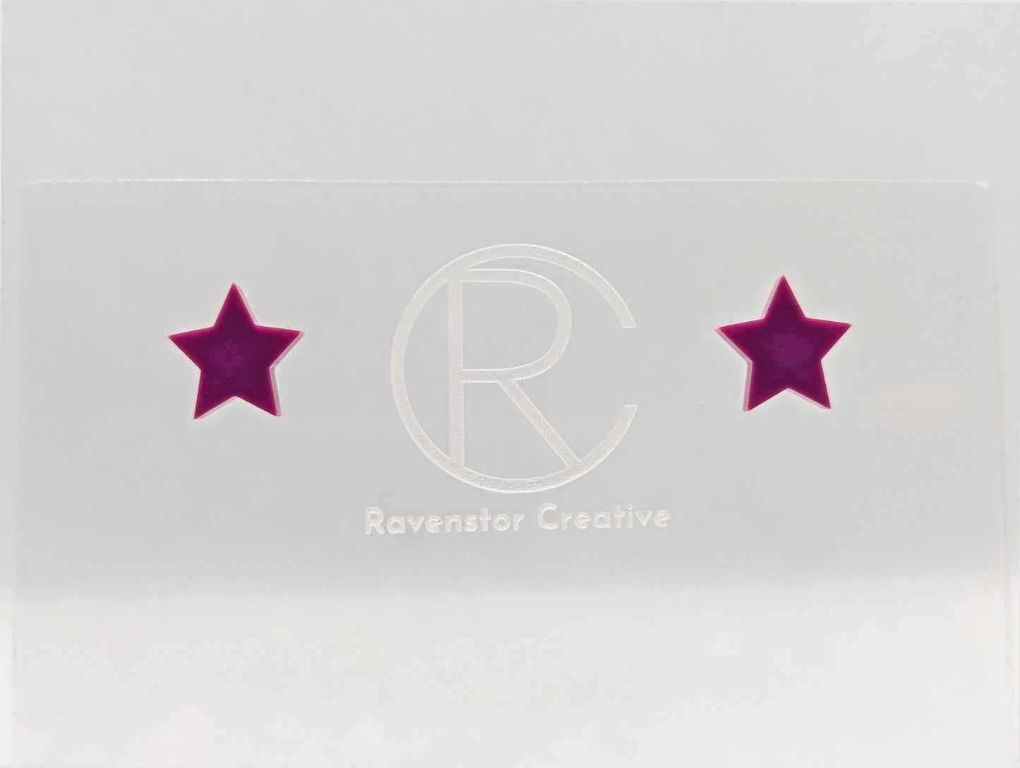 Shows a pair of small studded acrylic earrings that are Stars. They are Grape in colour. They are against a white background on see through stand with the ravenstor creative logo on.