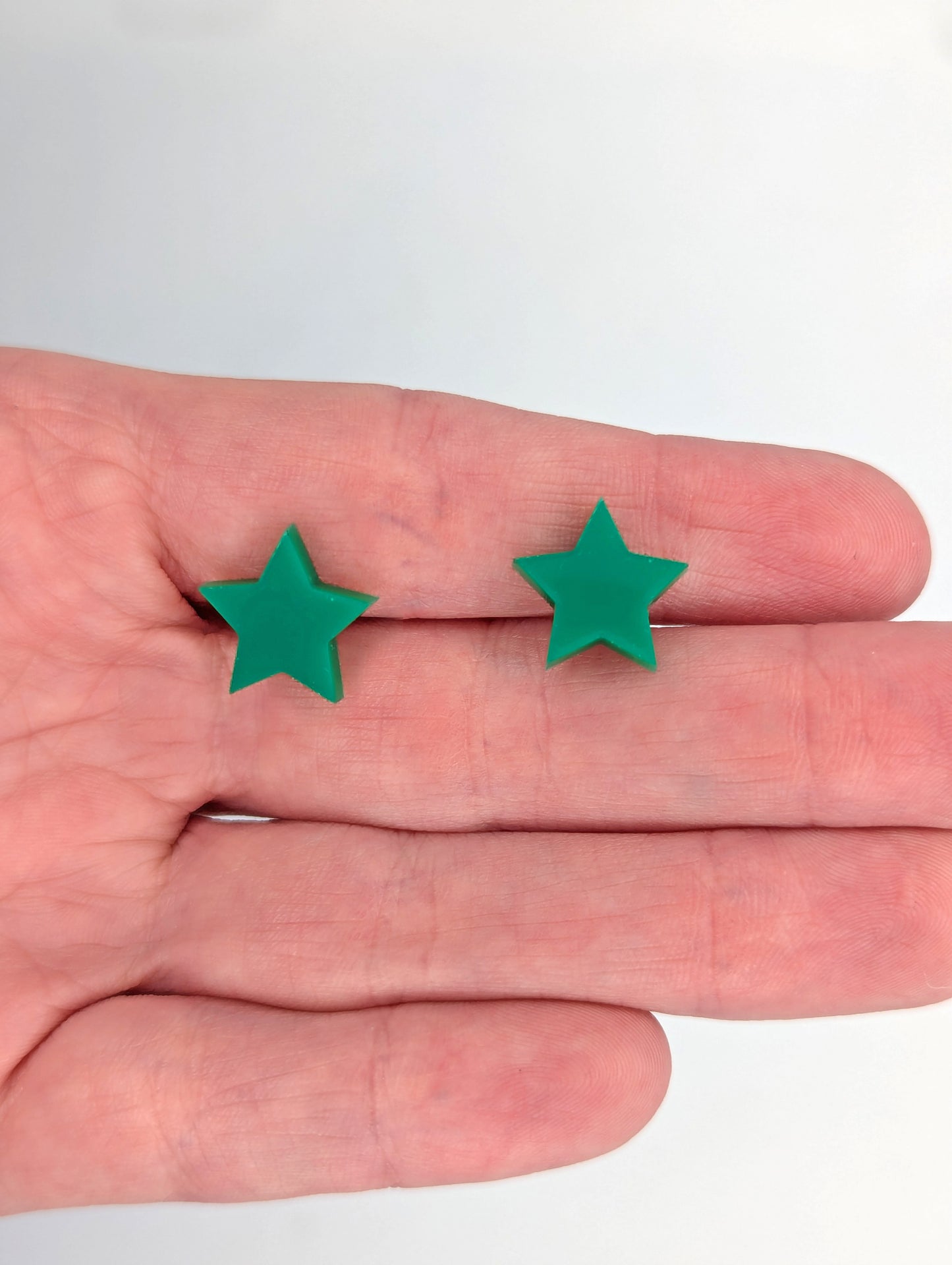 Shows a pair of small studded acrylic earrings that are love Hearts. The colour is Green. They are against a hand showing the size of one finger width in length and slightly smaller width.