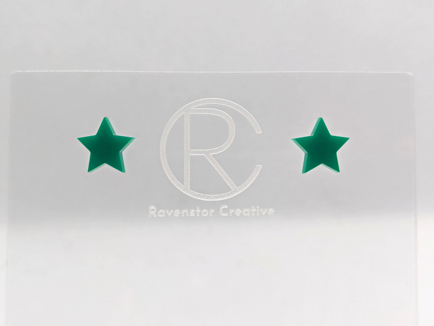 Shows a pair of small studded acrylic earrings that are Stars. They are Green in colour. They are against a white background on see through stand with the ravenstor creative logo on.