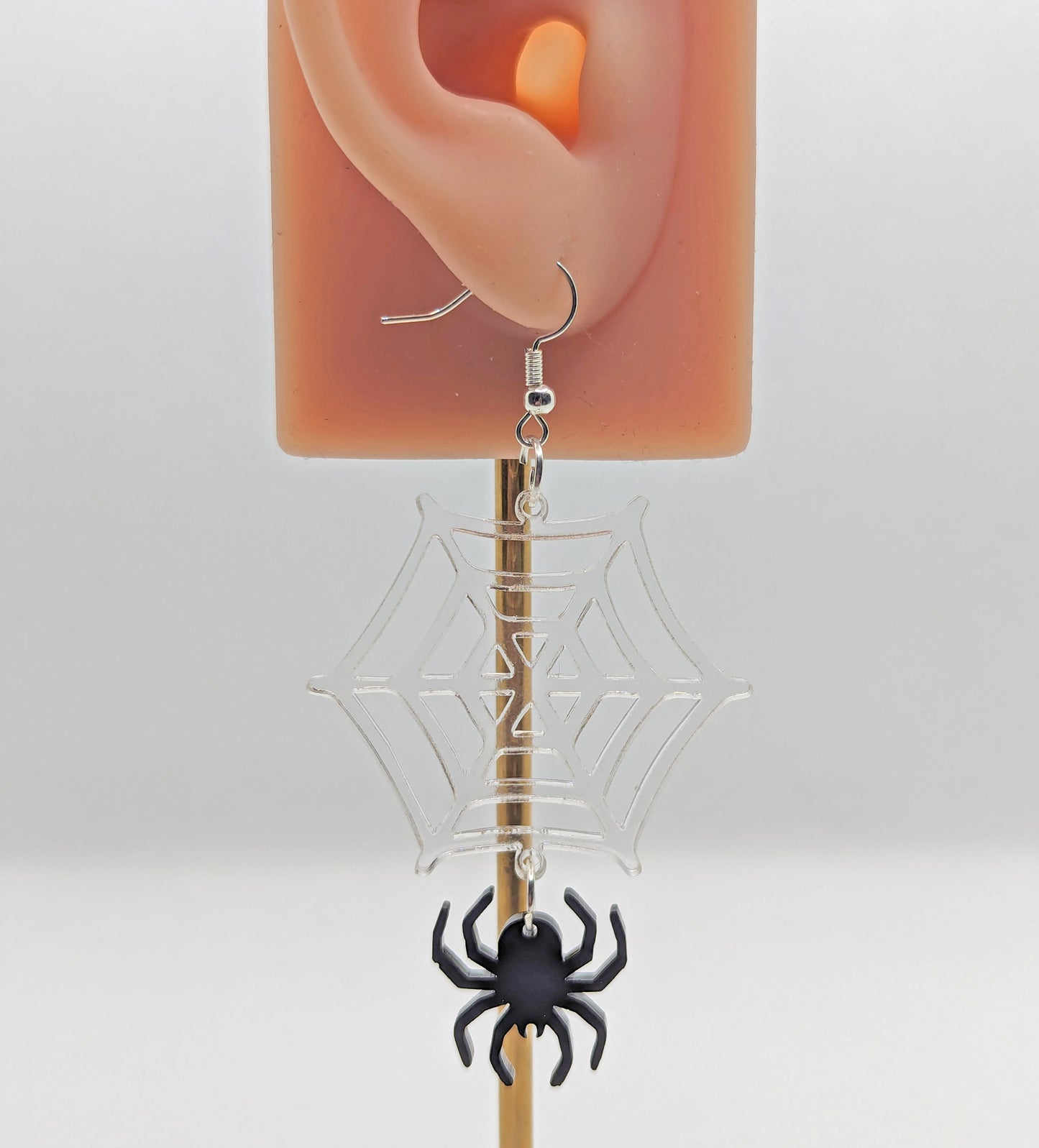 Image of a spider dangling from a web earring on an ear showing the size against a white background. The colours are a clear / see through web and black spider and have silver ear wire hook fixings.