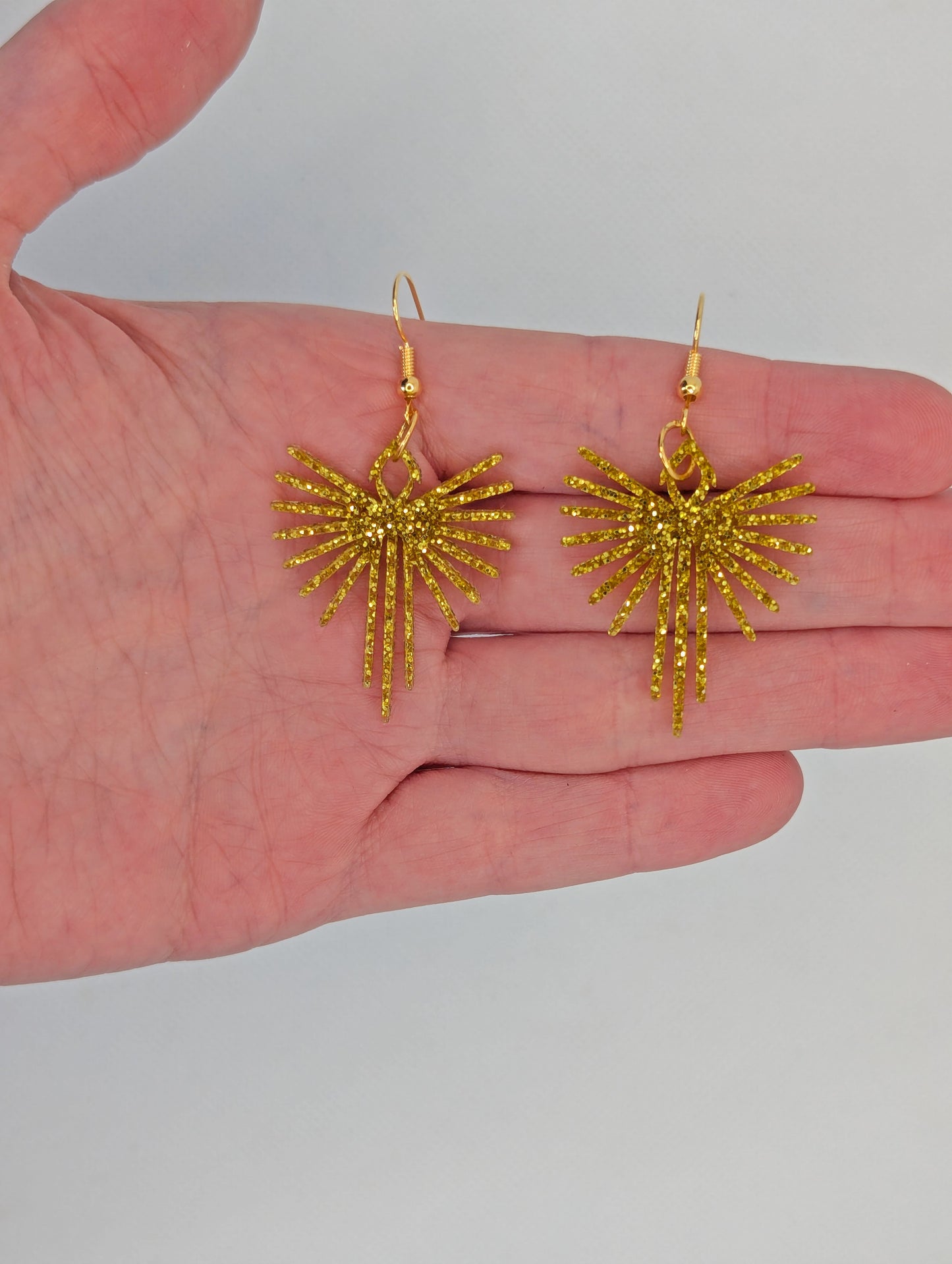 Shows a pair of earrings that have Gold ear wire hook fixings. They are Decorative Pointed Spread Wing earrings. The colour of them is Gold Glitter. They are against a white background against a hand to show the size of two finger widths in length and width.