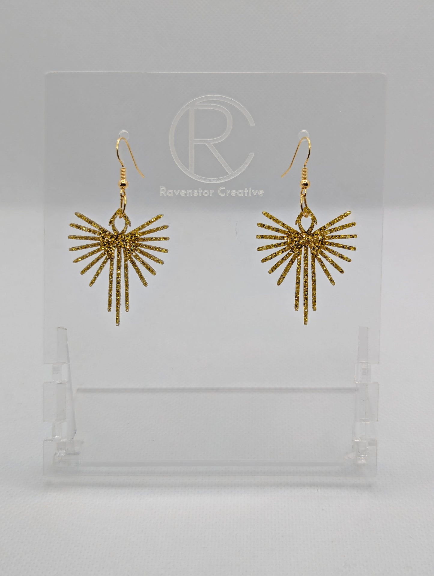 Shows a pair of earrings that have Gold ear wire hook fixings. They are Decorative Pointed Spread Wing earrings. The colour of them is Gold Glitter. They are against a white background on see through stands with the Ravenstor Creative logo.