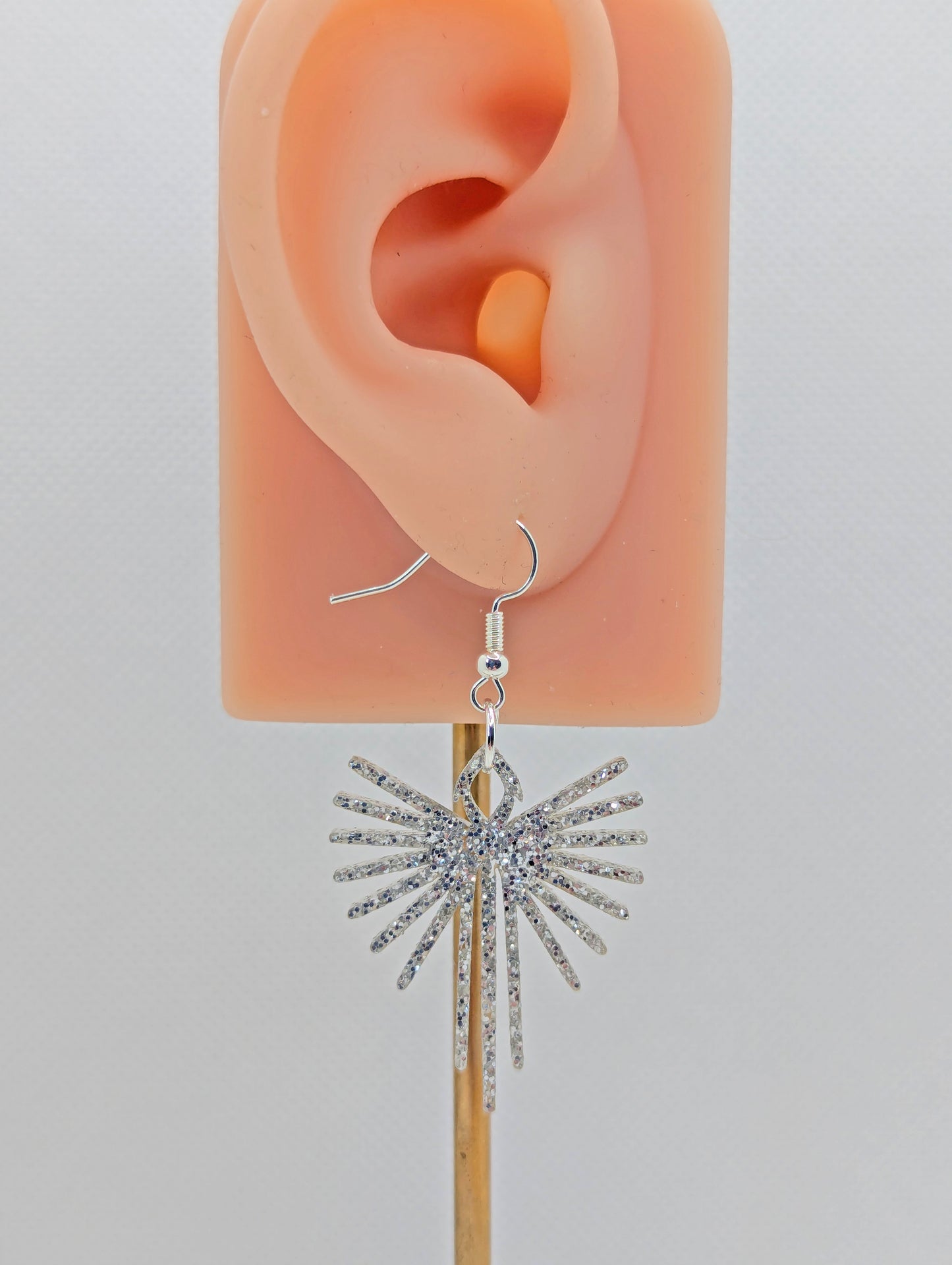 Shows an earring that has a Silver ear wire hook fixing. it is a Decorative Pointed Spread Wing earring. The colour of them is Silver Glitter. They are on an ear to show the size against a white background.