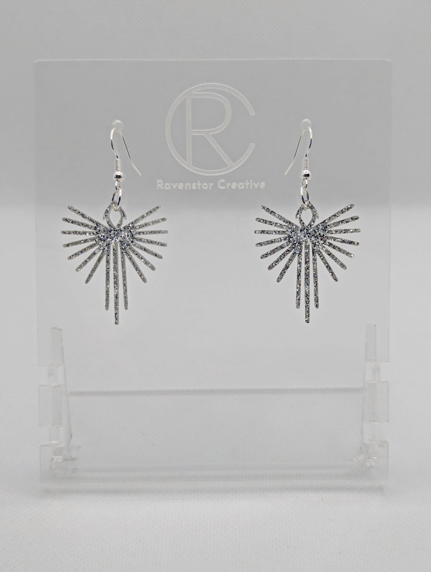 Shows a pair of earrings that have Silver ear wire hook fixings. They are Decorative Pointed Spread Wing earrings. The colour of them is Silver Glitter. They are against a white background on see through stands with the Ravenstor Creative logo.