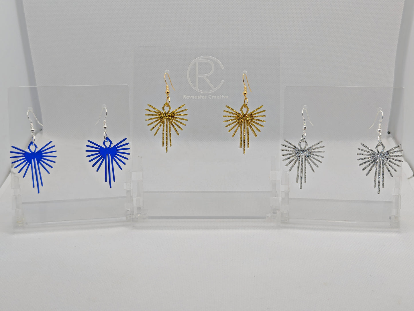 Shows three pairs of earrings that have ear wire hook fixings. They are Decorative Pointed Wing earrings. The colours are Translucent Blue, gold glitter and silver glitter. They are against a white background on see through stands with the Ravenstor Creative logo.