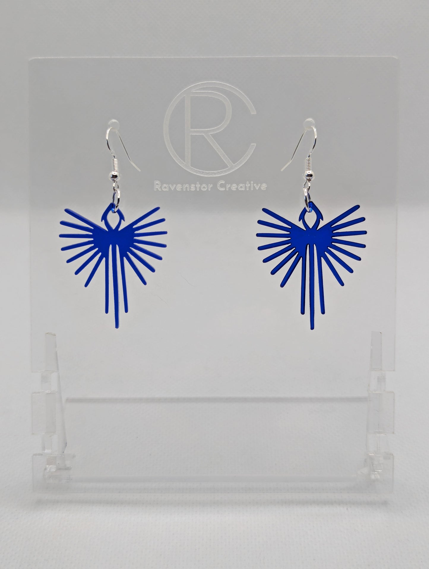 Shows a pair of earrings that have Silver ear wire hook fixings. They are Decorative Pointed Spread Wing earrings. The colour of them is Translucent Blue. They are against a white background on see through stands with the Ravenstor Creative logo.