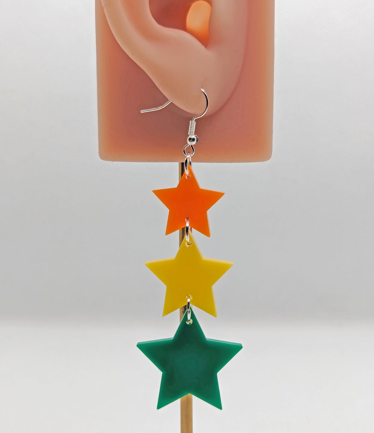 A dangly earring, which has three stars dangling from each other, the top is the smallest and they get bigger as they go down. It is on an ear to show the size. They are made from Orange, Yellow and Green Acrylic with silver ear wire hook fixings.