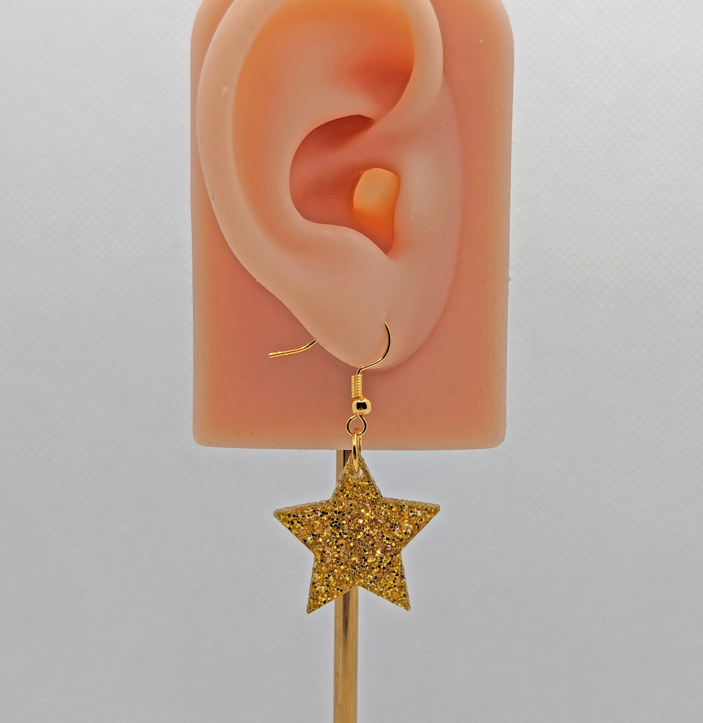 Image of a pair of five pointed star shaped earrings on an ear showing the size against a white background. They are gold glitter and have gold fixings.