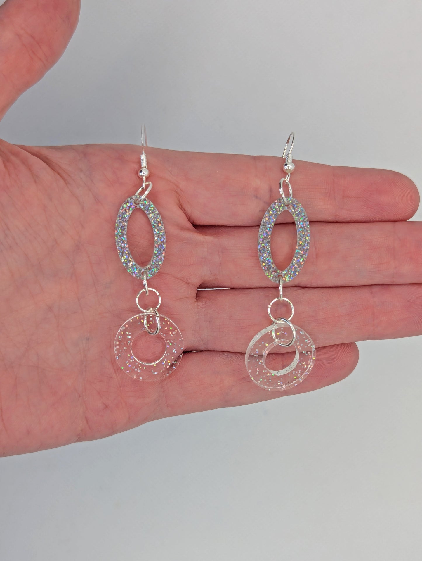 A two part drop earring with a Silver ear wire hook fixing. It is an oval with a circle dangling underneath joined by three Silver jump rings. It is against a hand to show the size of four finger widths in length and one and a half wide.