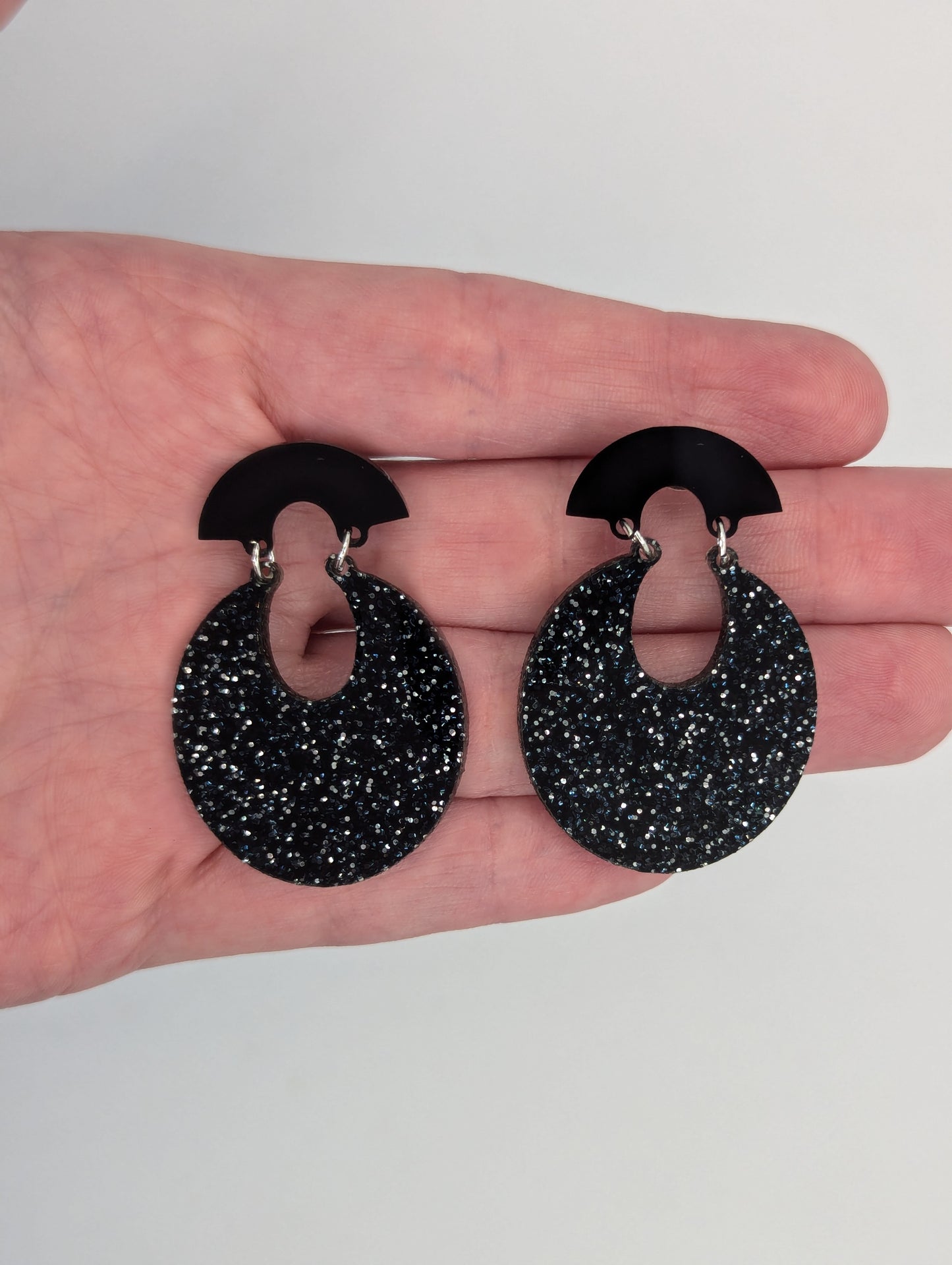 A pair of acrylic stud earrings. They are a small fan shape with an open hoop dangling underneath. They are against a hand to show the size of Three finger widths in length and two wide.