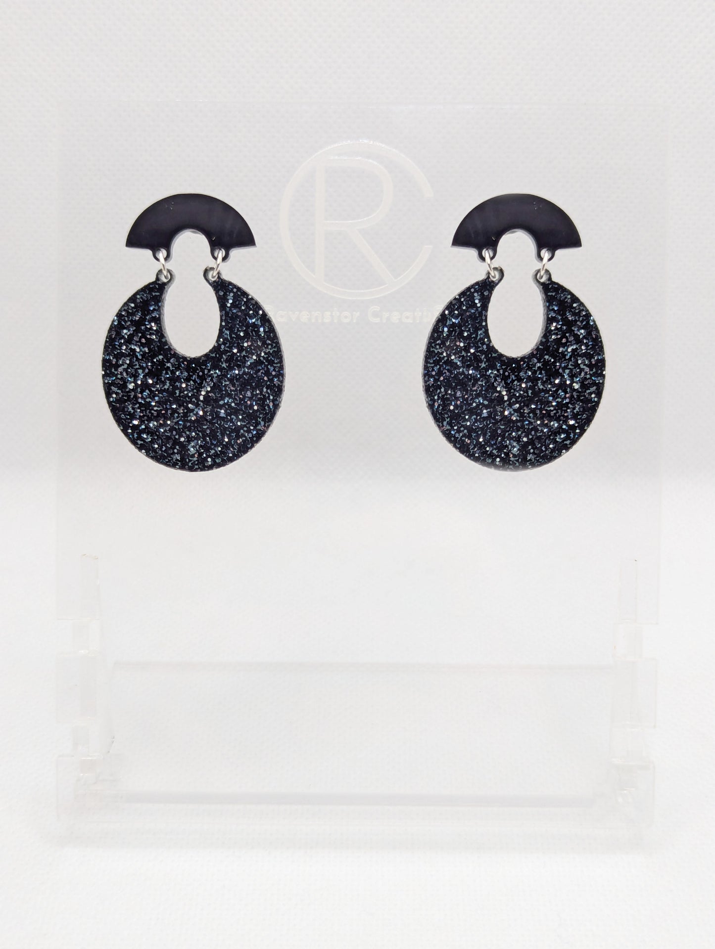 A pair of acrylic stud earrings. They are a small fan shape with an open hoop dangling underneath. The top fan is Black and bottom part is an open hoop shape and is Silver and Black Glitter. They are on a clear stand against a white background.