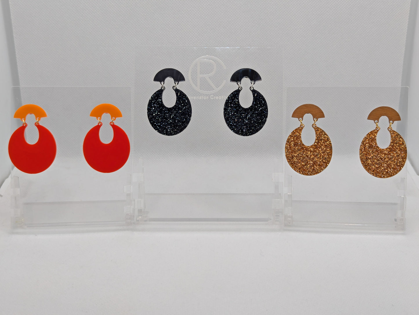 Three pairs of acrylic stud earrings. They are a small fan shape with an open hoop dangling underneath. The colours are Orange and Red, Black and Silver Glitter, Cherry Wood and Copper Glitter. They are on a clear stand against a white background.