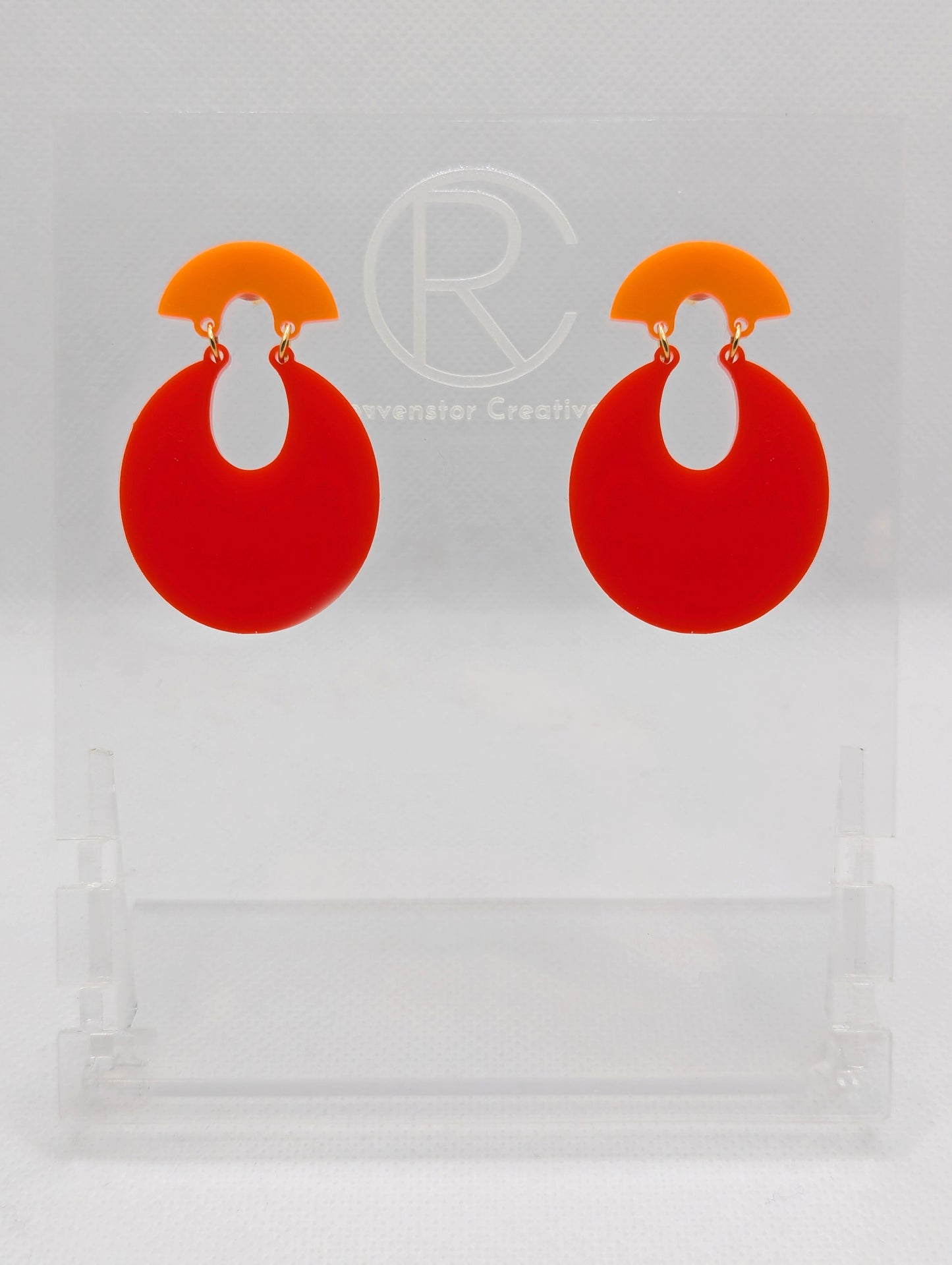 A pair of acrylic stud earrings. They are a small fan shape with an open hoop dangling underneath. The top fan is Orange and bottom part is an open hoop shape and is Red. They are on a clear stand against a white background.