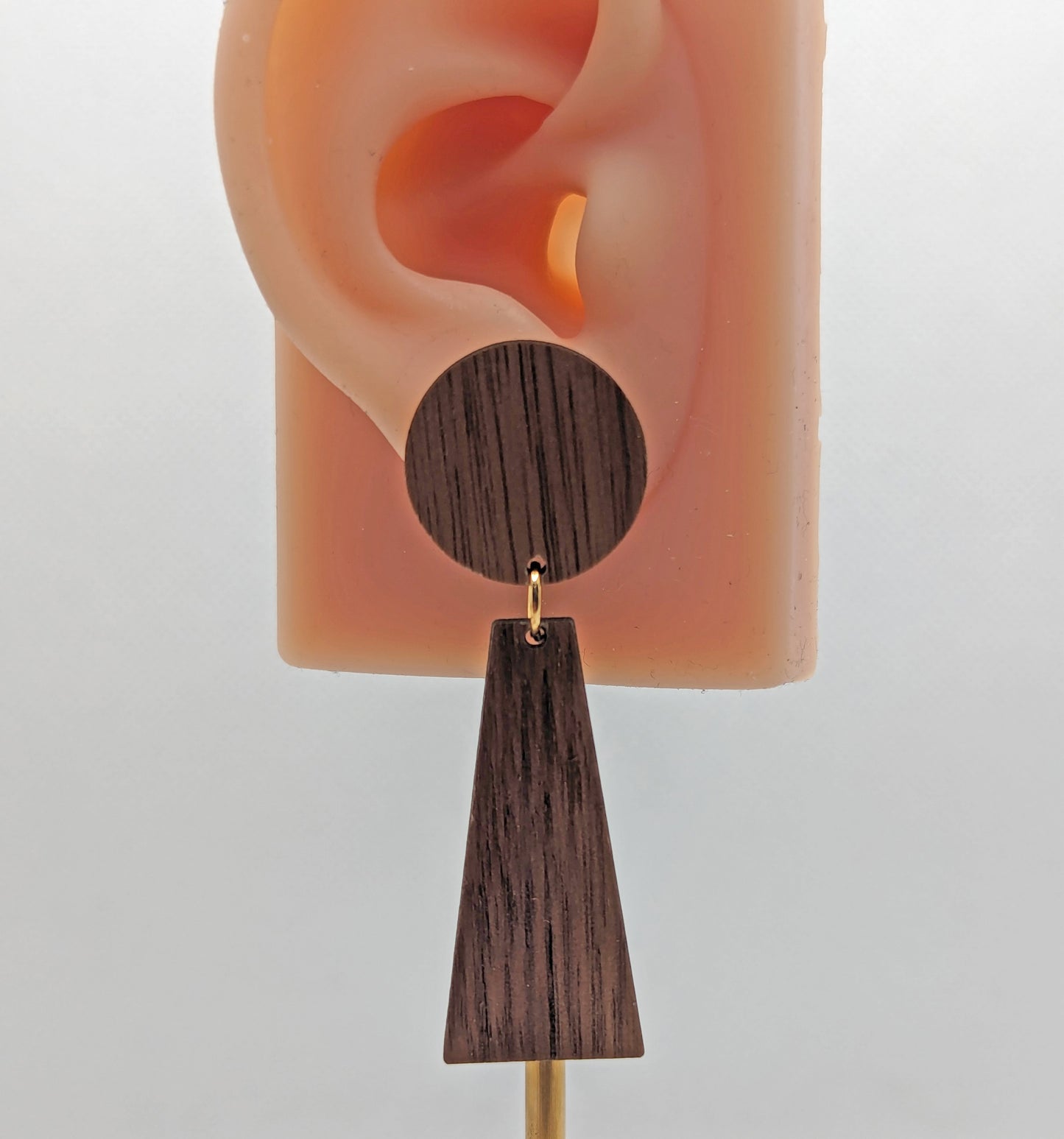 Shows an earring that is two parts on an ear to show the size. Stud fixings are attached to a circle, dangling under is a trapezoid. It is made from walnut wood with gold fixings.