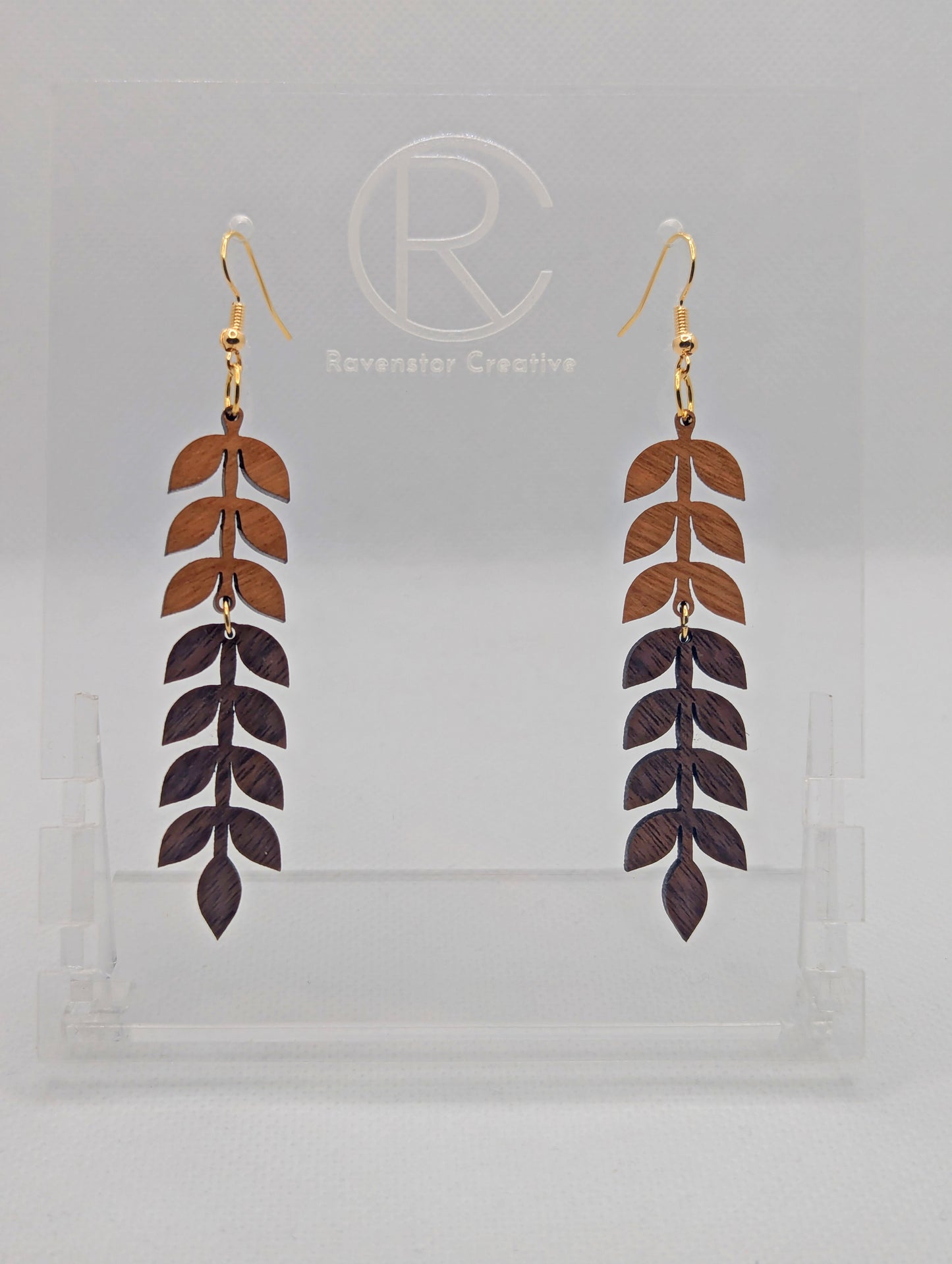 An image of two part wooden drop earrings with Gold ear wire hook fixings. The top part is Cherry Wood and the Bottom part is Walnut Wood. They are on a clear stand with the Ravenstor Creative logo on it against a white background.
