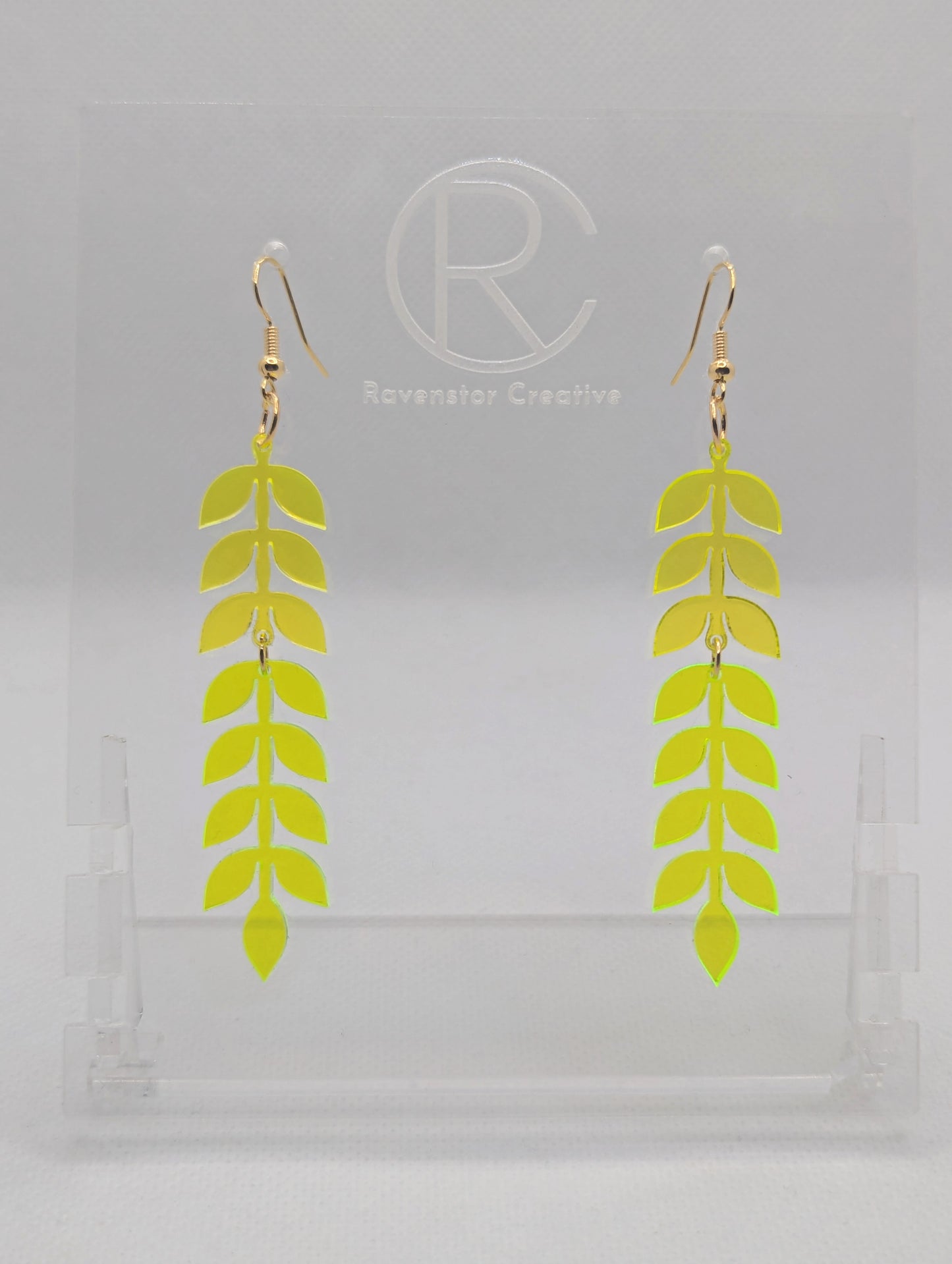 An image of two part wooden drop earrings with Gold ear wire hook fixings. The top part is Neon Orange and the Bottom part is Neon Yellow. They are on a clear stand with the Ravenstor Creative logo on it against a white background.