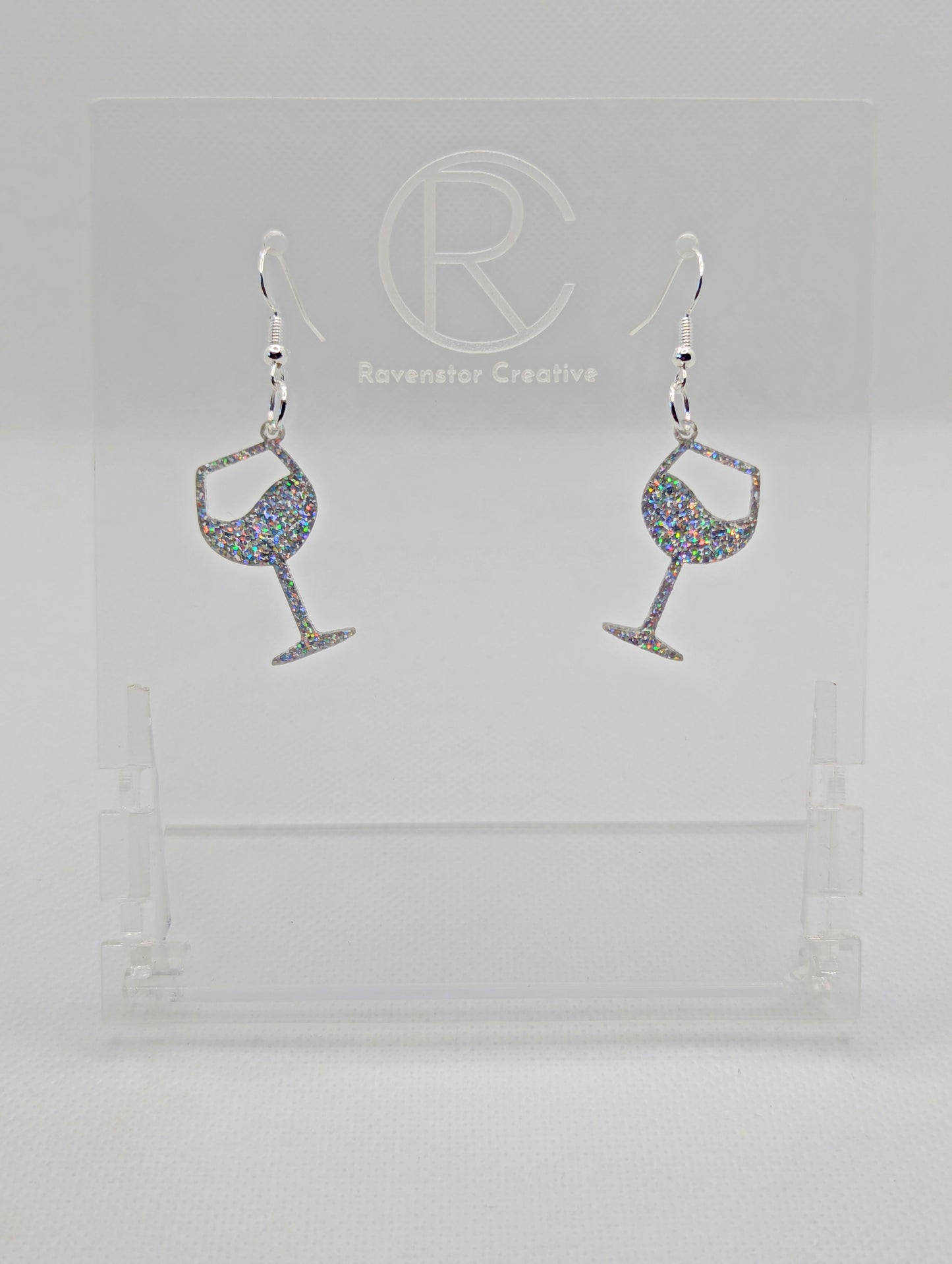 An image of a pair of Wine Glass Earrings with Silver ear wire hook fixings. These are ideal for a Hen Do or Bachelorette Party. The colour is Holographic Glitter. They are on clear stands against a white background.