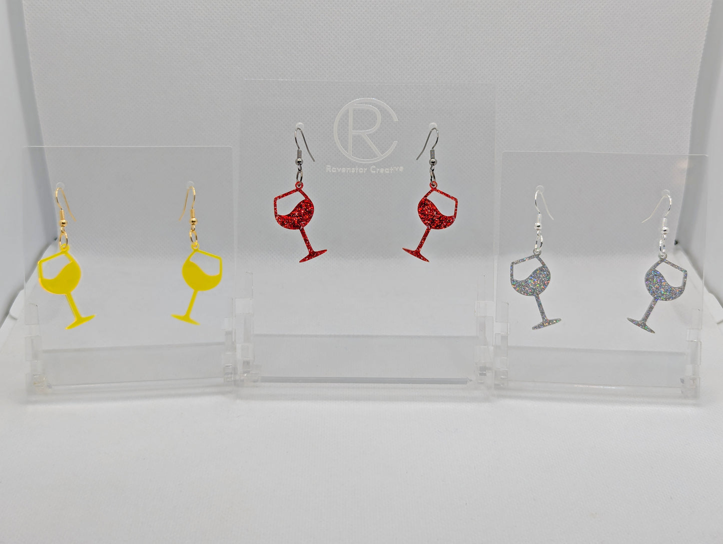 An image of three pairs of Wine Glass Earrings with ear wire hook fixings. These are ideal for a Hen Do or Bachelorette Party. The colours are Neon Orange, Red Glitter and Holographic Glitter. They are on clear stands against a white background
