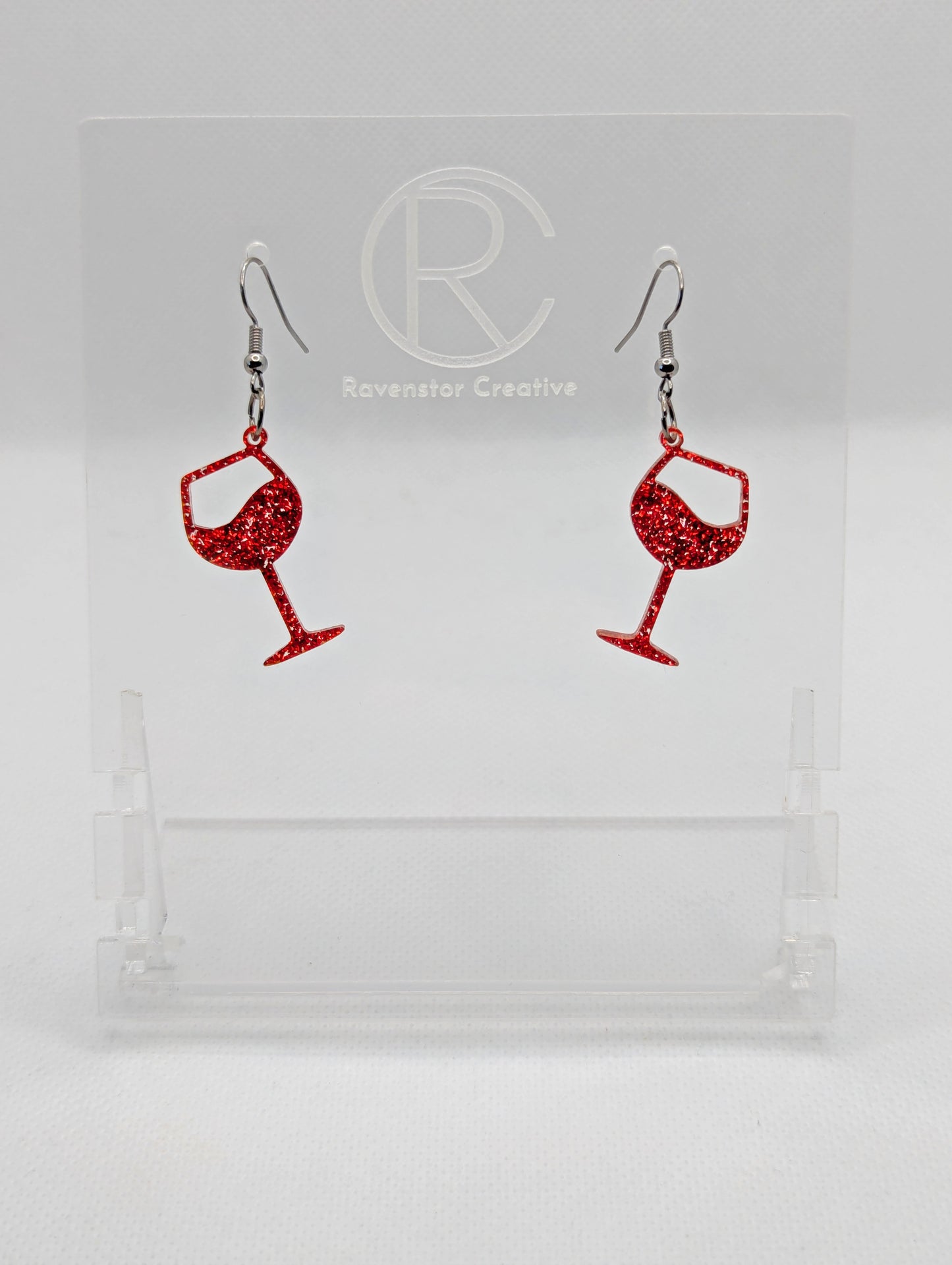 An image of a pair of Wine Glass Earrings with Black ear wire hook fixings. These are ideal for a Hen Do or Bachelorette Party. The colour is Red Glitter. They are on clear stands against a white background.
