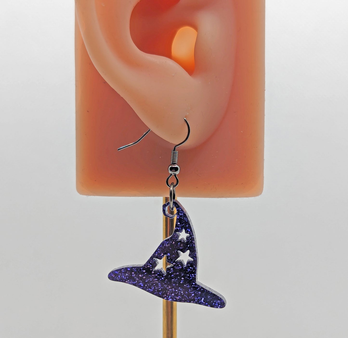Image of a Halloween Witches Hat shape earring with stars cut out on an ear showing the size against a white background. It is made from purple glitter acrylic and have black ear wire hook fixings.