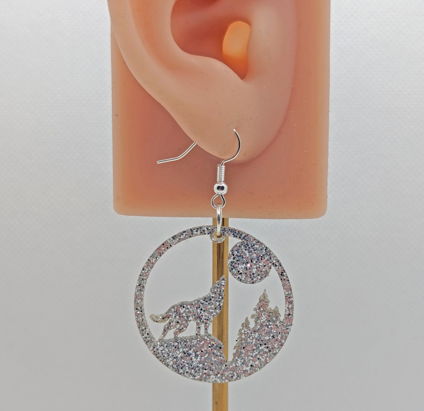 Image of a circle earring with a wolf howling at the moon cut out, on an ear showing the size against a white background. It is made from Silver Glitter and have silver ear wire hook fixings.