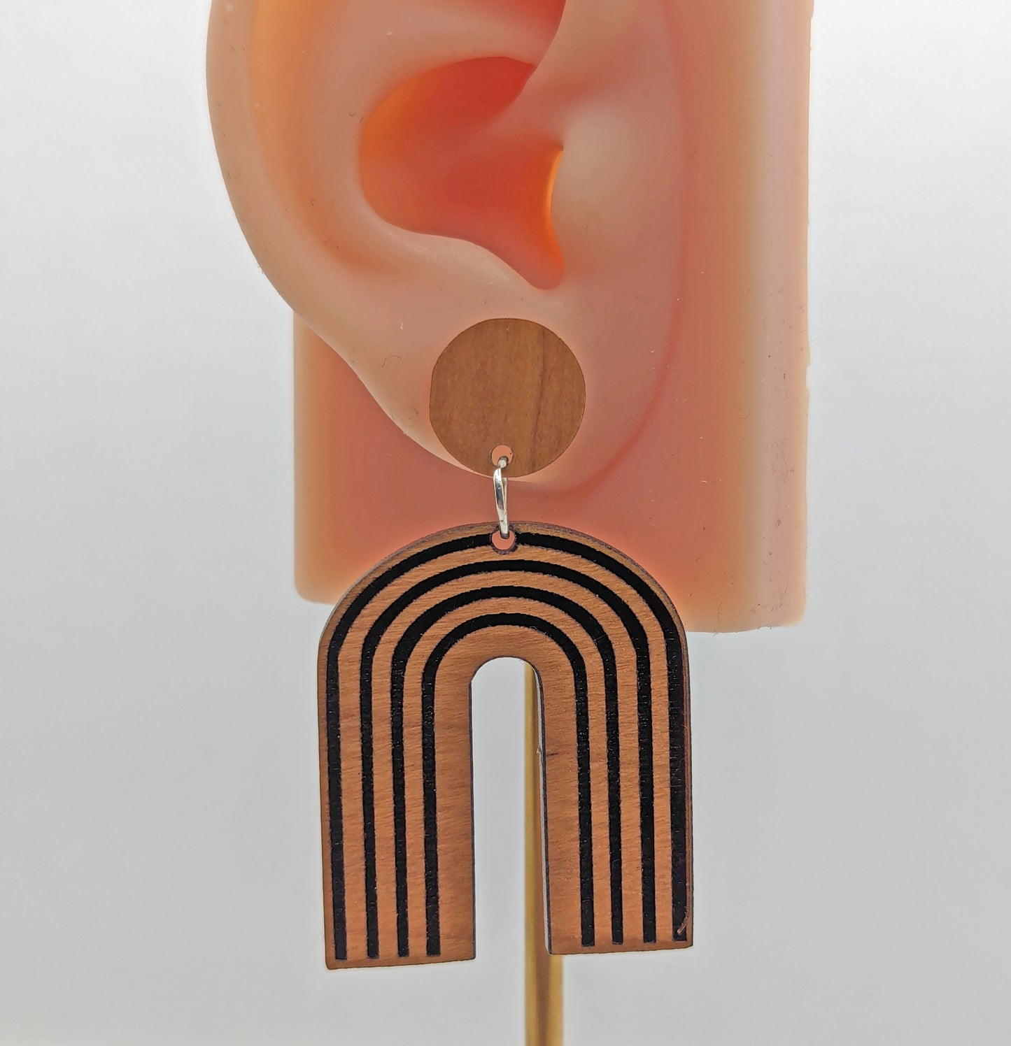 An arch earring dangling under a circle with stud fixings. Engraved are lines like a wooden rainbow 80s retro style. It is made from cherry wood and are on an ear to show the size against a white background.