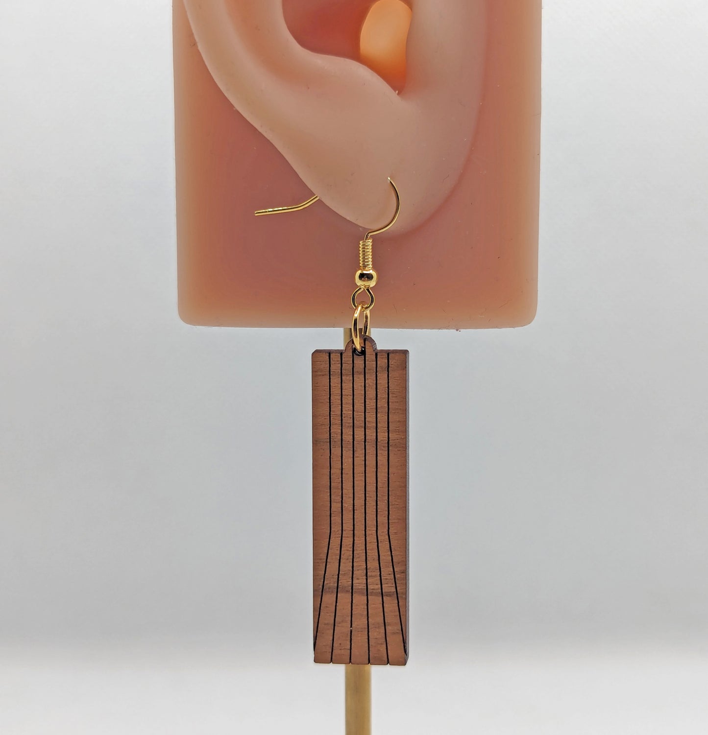 An earring on an ear showing the size against a white background. It is a slim rectangle with vertical lines running from the top to the bottom that fan out. It has gold ear wire hook earrings and are against a white background.