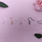 An image of the different fixings these earrings are available with. They are silver, Gold, Rose Gold, Black and a Silver Clip-on clasp. They are displayed on a light pink / purple background with some leaves and rose head in the corners.