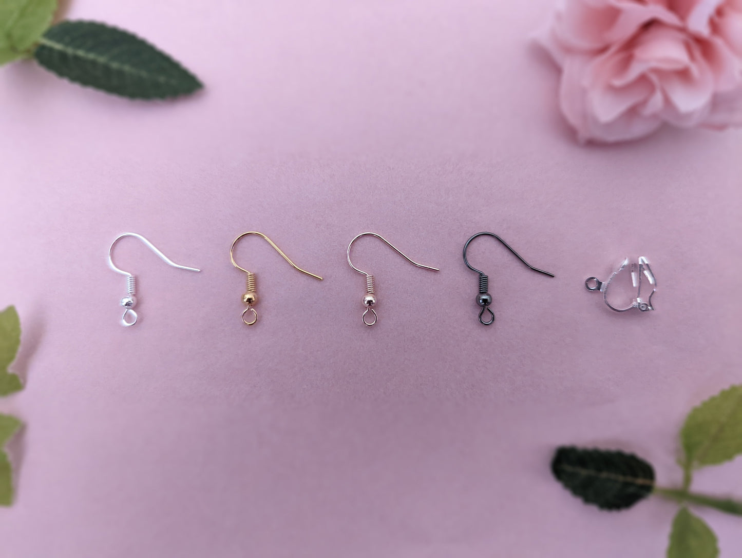 An image of the different fixings these earrings are available with. They are silver, Gold, Rose Gold, Black and a Silver Clip-on clasp. They are displayed on a light pink / purple background with some leaves and rose head in the corners.