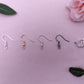 an image of the different fixings these earrings are available with. They are silver, Gold, Rose Gold, Black and a Silver Clip-on clasp. They are displayed on a light pink / purple background with some leaves and rose head in the corners.