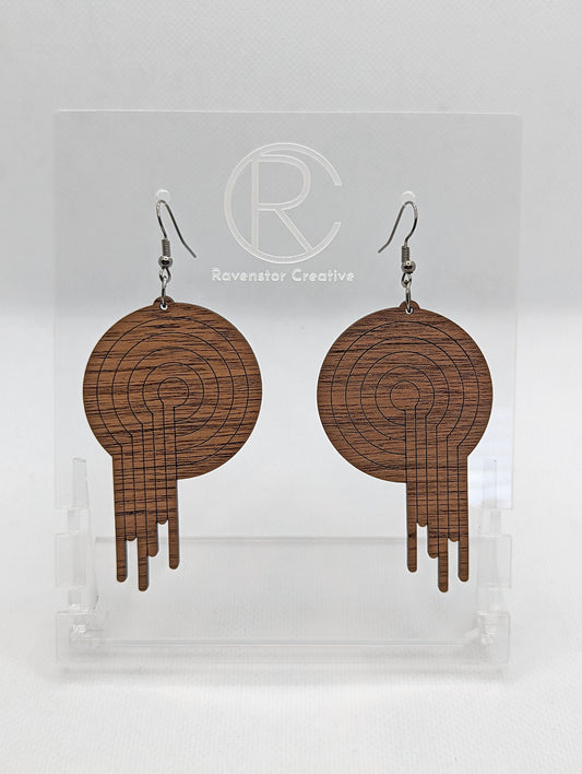 A pair of circle earrings with black ear wire hook fixings. Engraved in the centre is an 80s Retro melting record. They are made from walnut wood. They are against a white background on a clear stand with the Ravenstor Creative logo on it.