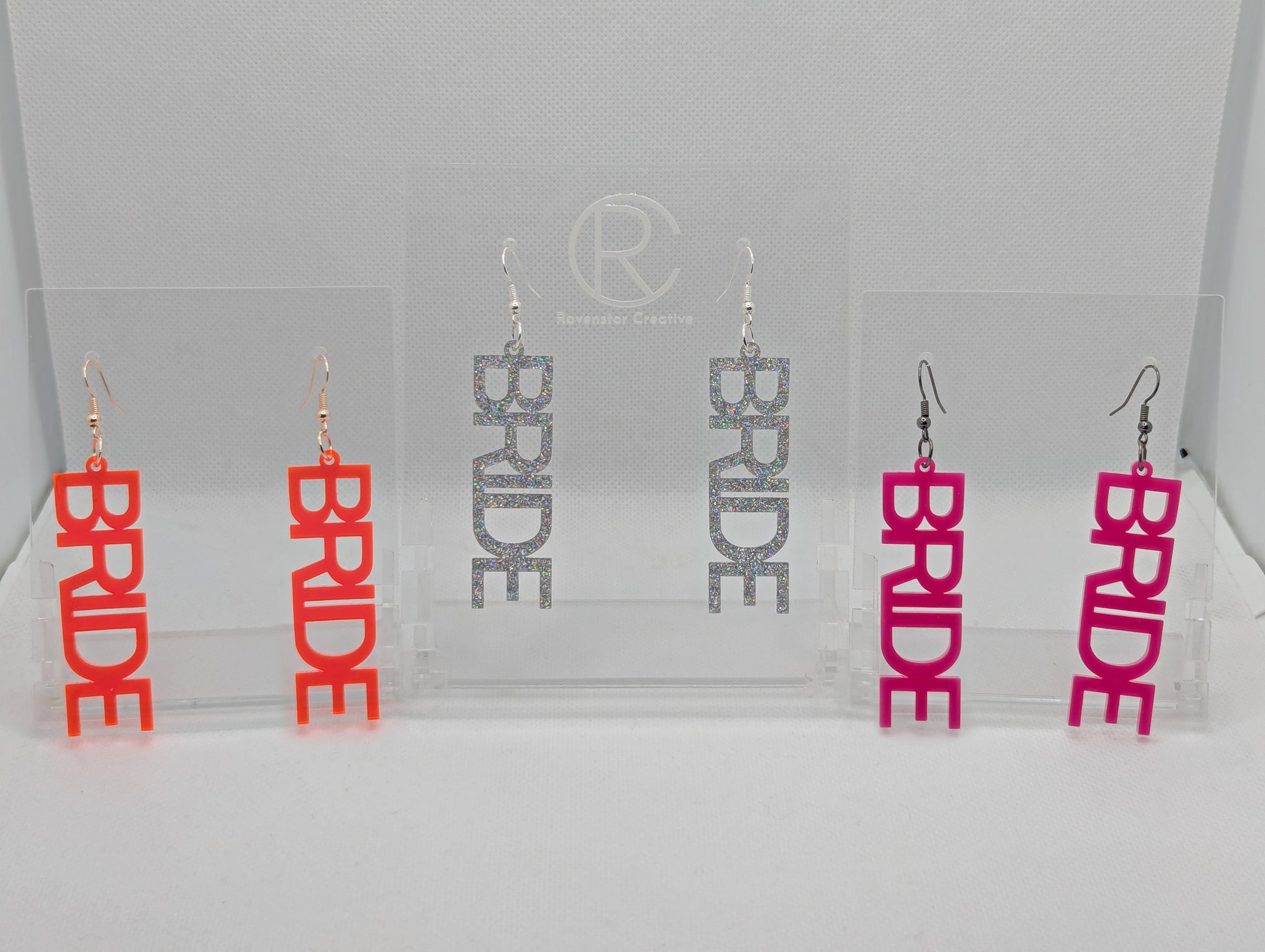 Image of Three pairs of earrings with ear wire hook fixings. Each one is the word Bride in capitals. The colours are Neon Pin, Holographic Glitter and Hot Pink. They are on clear stands against a white background.