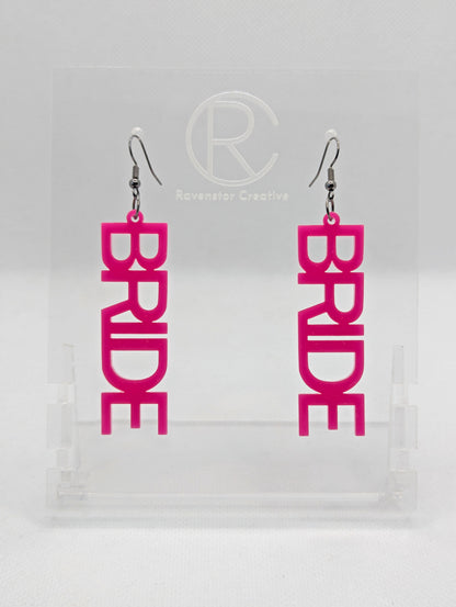 A pair of earrings with Black ear wire hook fixings. Each one is the word Bride in capital letters. They are Hot Pink in colour. These are ideal for a Hen do or Bachelorette Party. They are on clear stands against a white background.