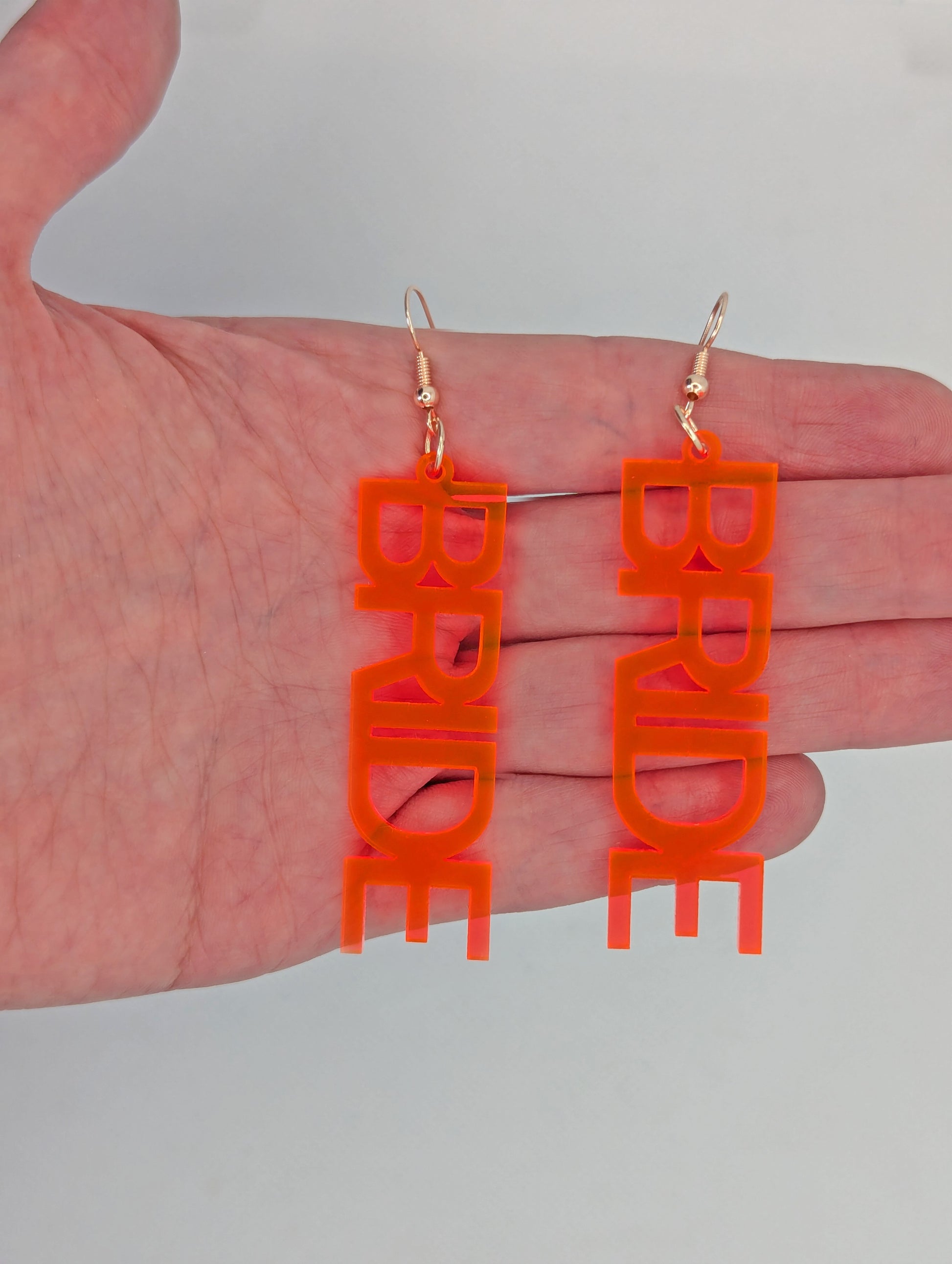 Earrings with Rose Gold ear wire hook fixings. Each one is the word Bride in capital letters. These are ideal for a Hen do or Bachelorette Party. They are against a hand to show the size of four finger widths in length and one and a half wide.