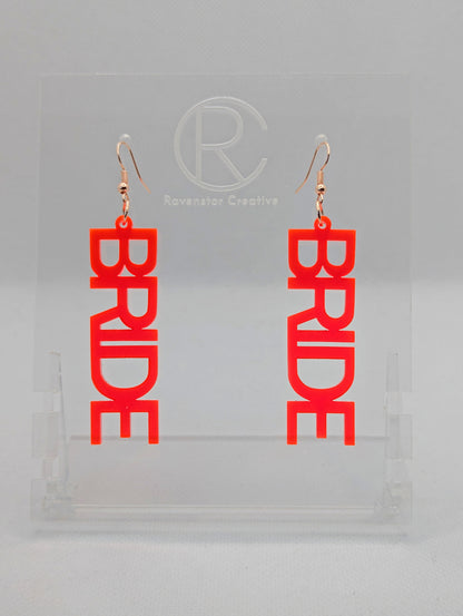 A pair of earrings with Rose Gold ear wire hook fixings. Each one is the word Bride in capital letters. They are Neon Pink in colour. These are ideal for a Hen do or Bachelorette Party. They are on clear stands against a white background.
