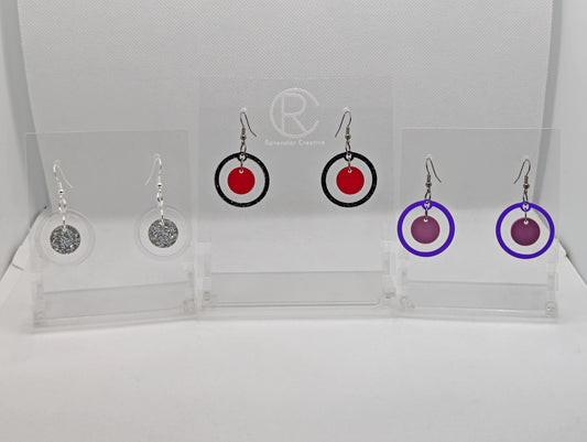 Shows three pairs earrings ear wire hook fixings. They are circle shapes with a smaller circle dangling inside them. The colours are Clear outer with Silver glitter, Black & Silver outer, translucent red inner and purple outer and Grape inner.