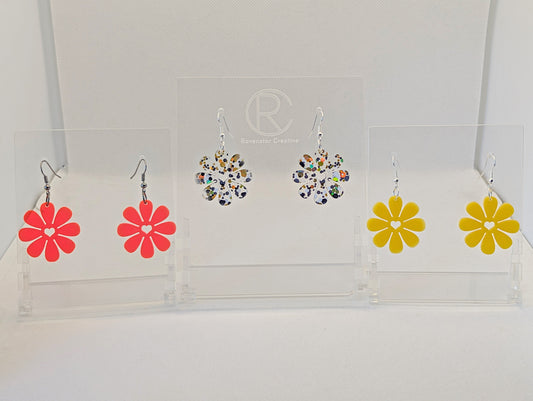 Shows three pairs of acrylic earrings that have ear wire fixings. They are Daisey Flower earrings with a heart cut out the the middle. The colours are Neon Pink, Lollipop Glitter and Yellow. They are against a white background on see through stand.