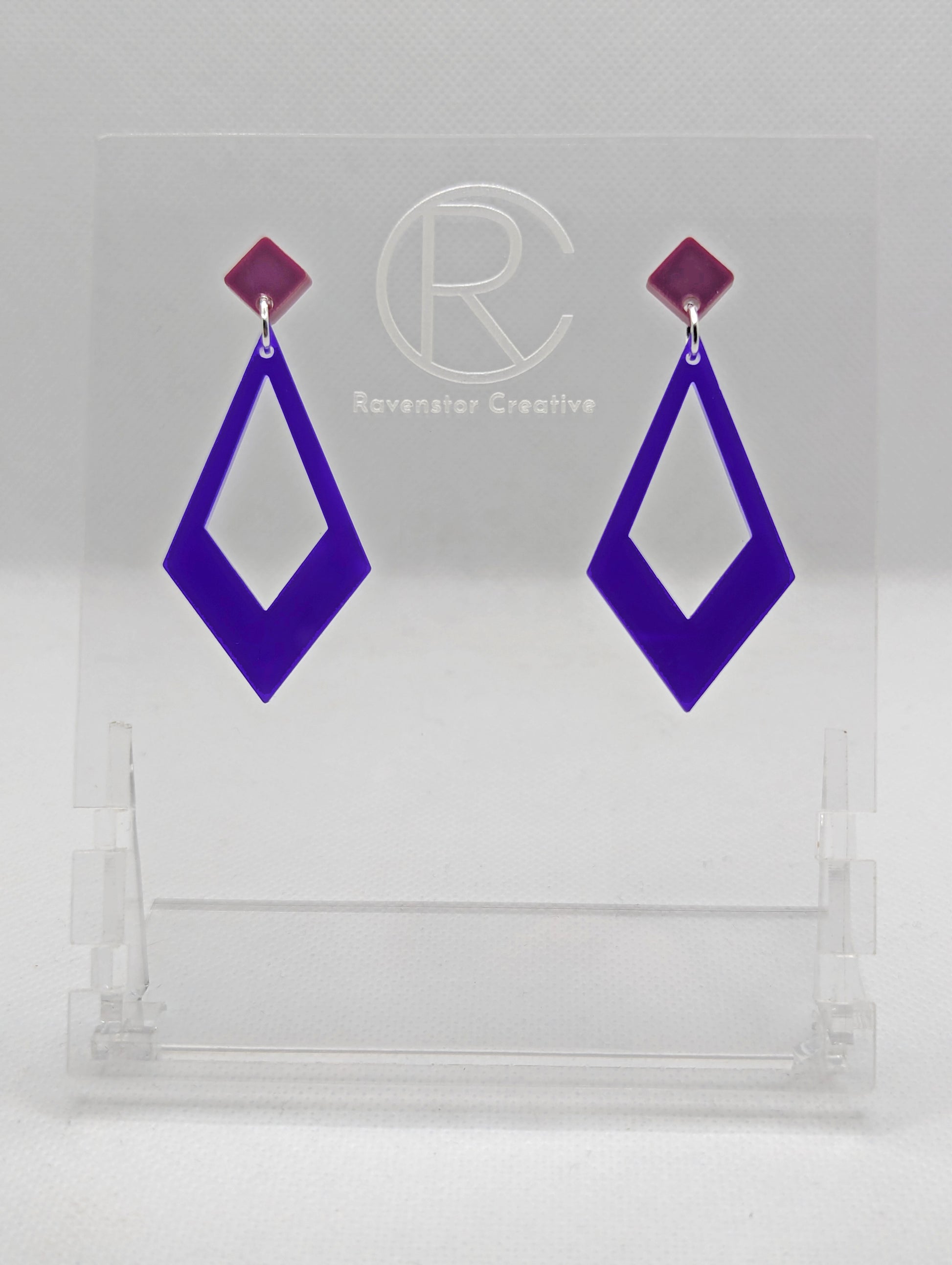 Shows a pair of 2 part earrings with stud fixings. The stud is on a small diamond shape with a larger kite shape dangling underneath. The colours are grape top and purple Kite. They are on a clear stand against a white backing