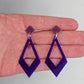 Shows a pair of 2 part earrings with stud fixings. The stud is on a small diamond shape with a larger kite shape dangling under on a hand showing thier size, they are three and a half finger widths in length and two wide. Colours are grape and purple