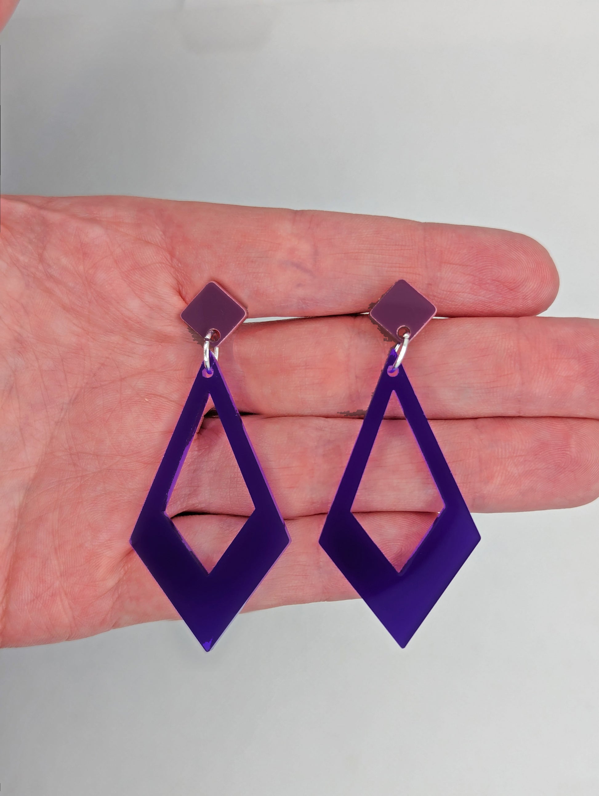 Shows a pair of 2 part earrings with stud fixings. The stud is on a small diamond shape with a larger kite shape dangling under on a hand showing thier size, they are three and a half finger widths in length and two wide. Colours are grape and purple